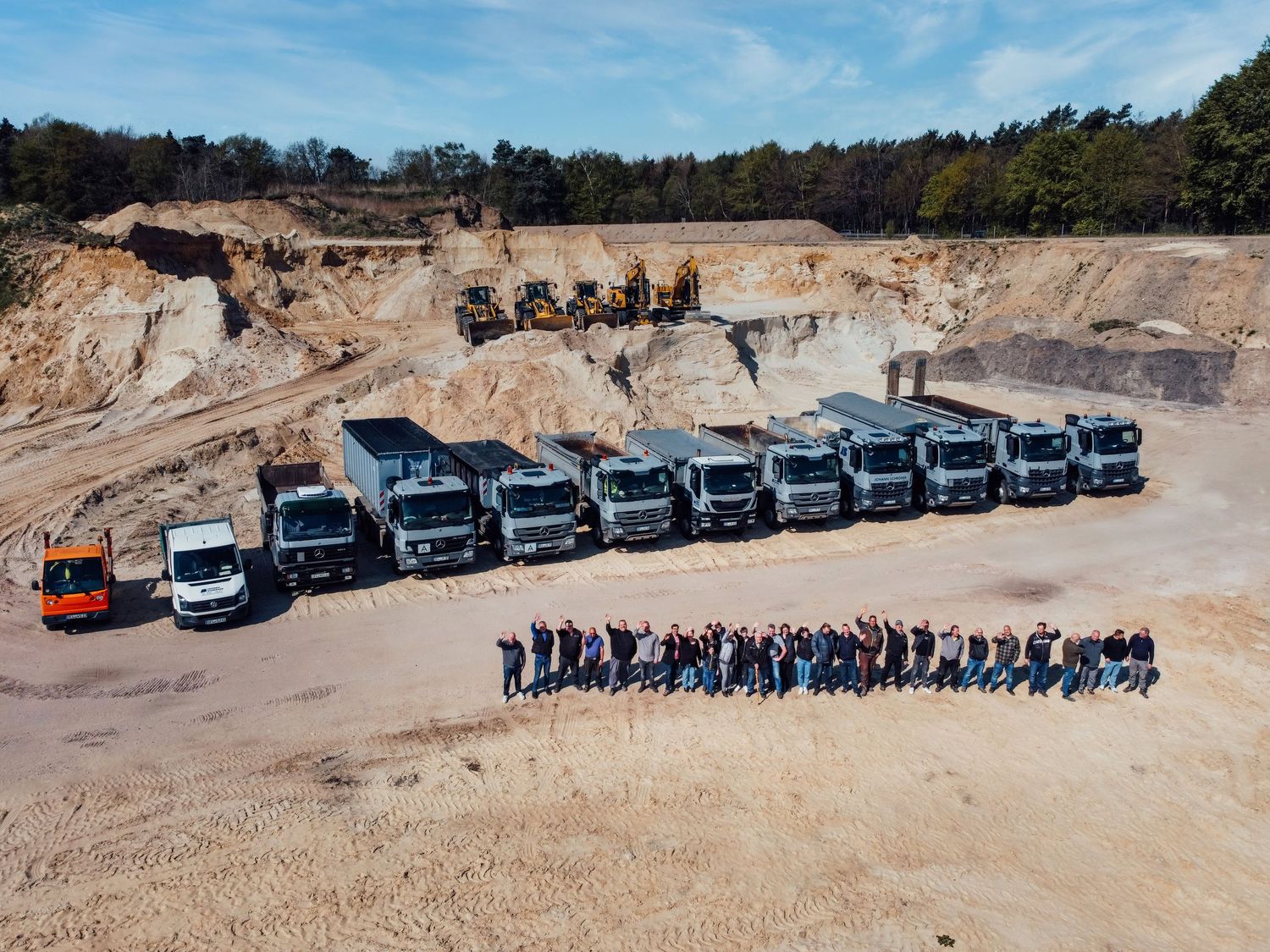 Aerial view of a fleet of dump trucks lined up in a quarry with workers posing in front.
