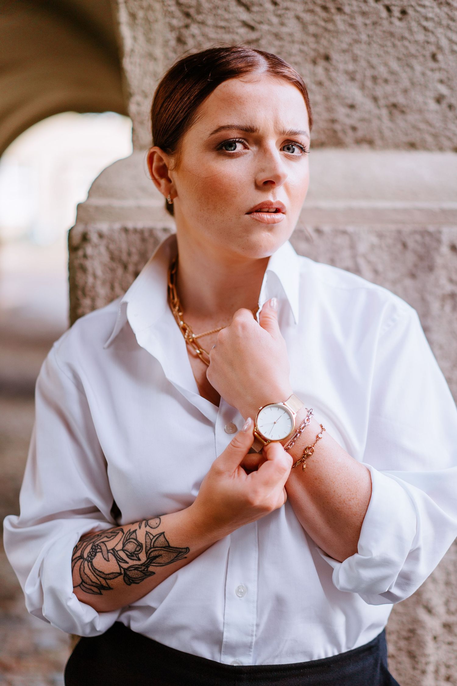 Young woman in white oversized shirt with tattoo and gold watch posing by stone column.