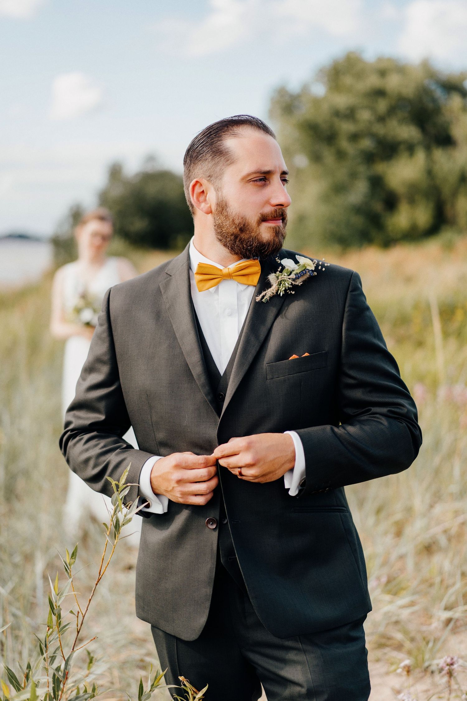 Bearded groom in dark suit with yellow bow tie adjusting jacket in field, bride visible in background.