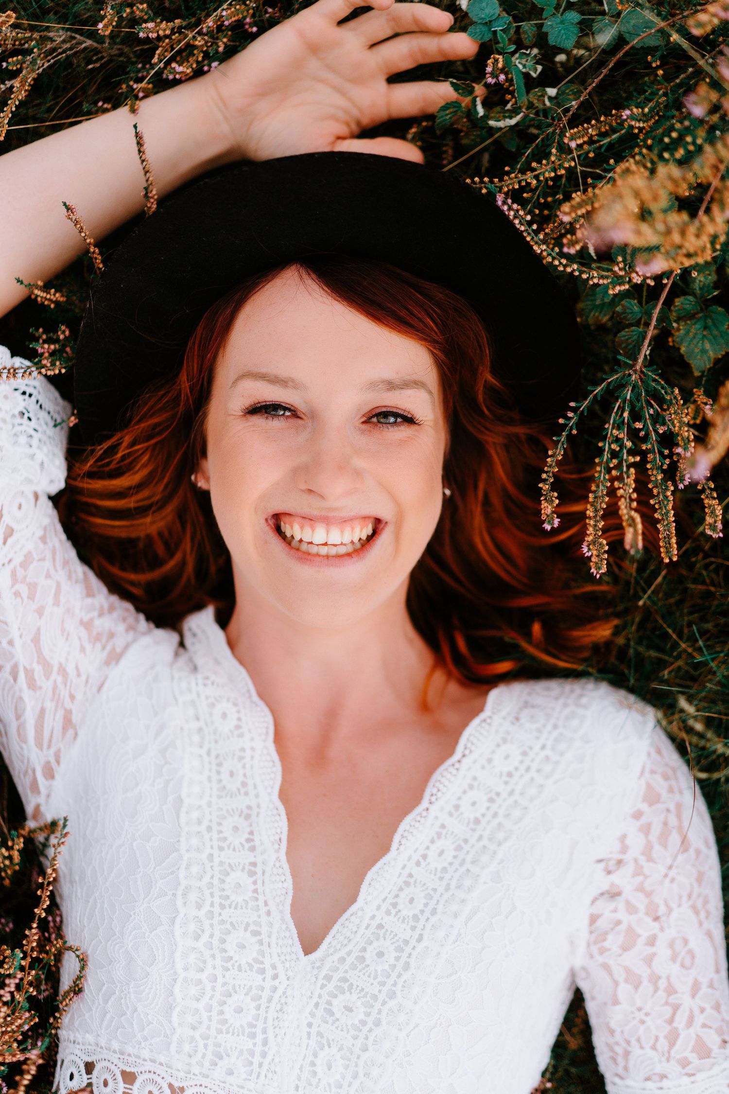 Smiling redhead woman lying in grass wearing white lace dress and black hat.