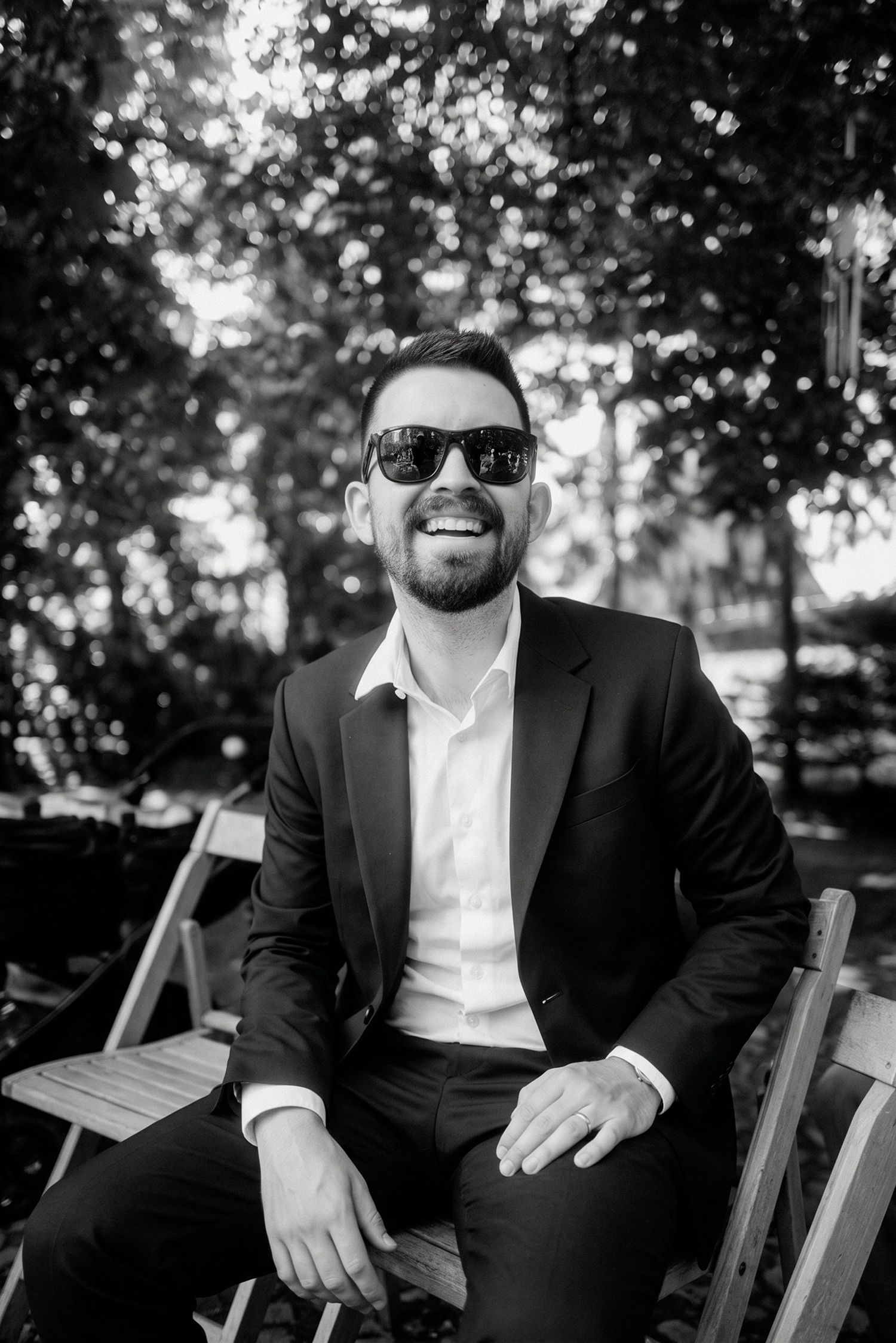 Smiling man in black suit and sunglasses sits outdoors among trees in black and white photo.