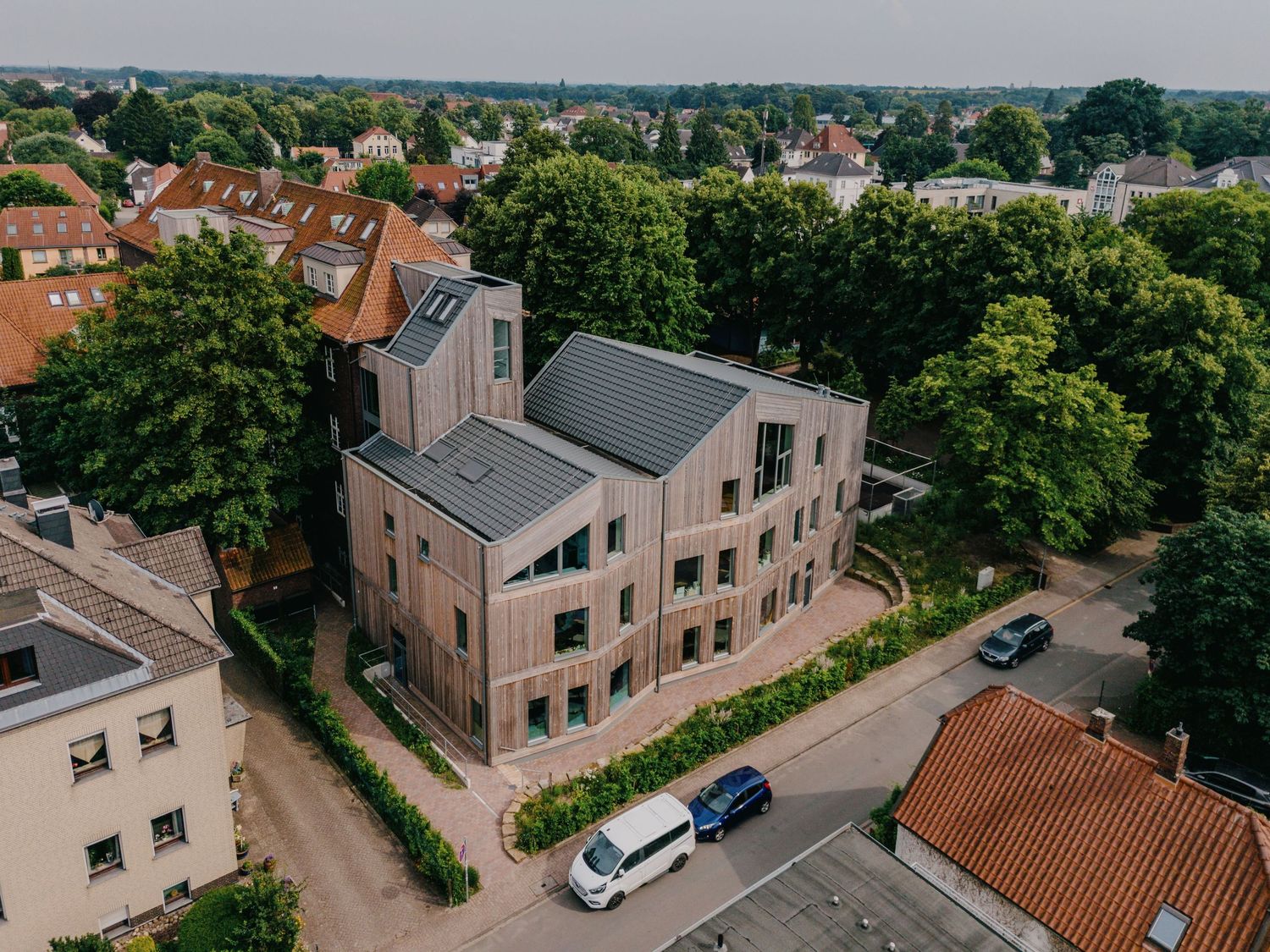 Aerial drone view of a modern brick and metal roof building surrounded by trees in a residential area.