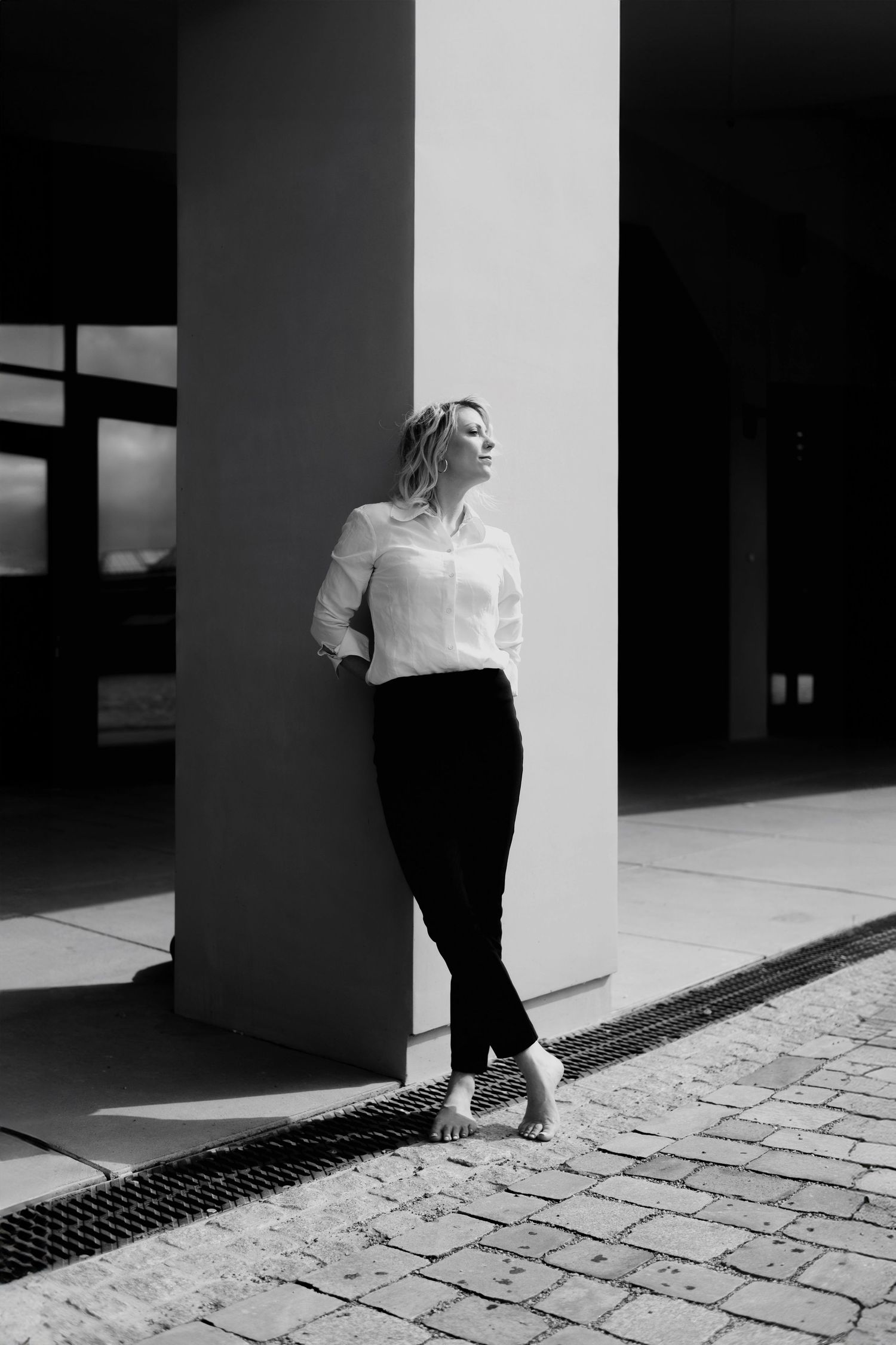 Black and white full-length portrait of blonde woman in white shirt leaning against pillar.