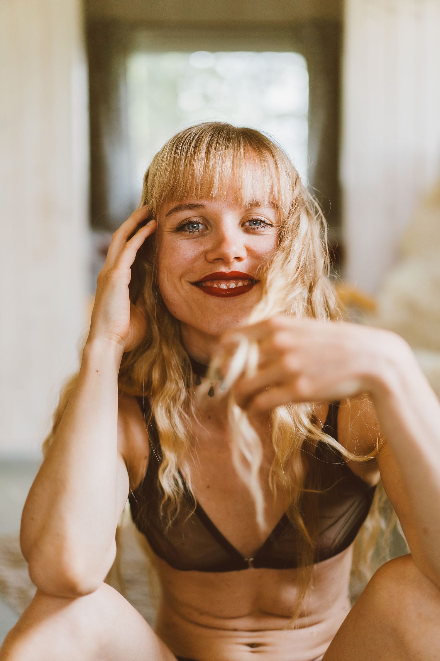 Smiling blonde woman in black sheer bra with red lips poses in bright indoor setting.