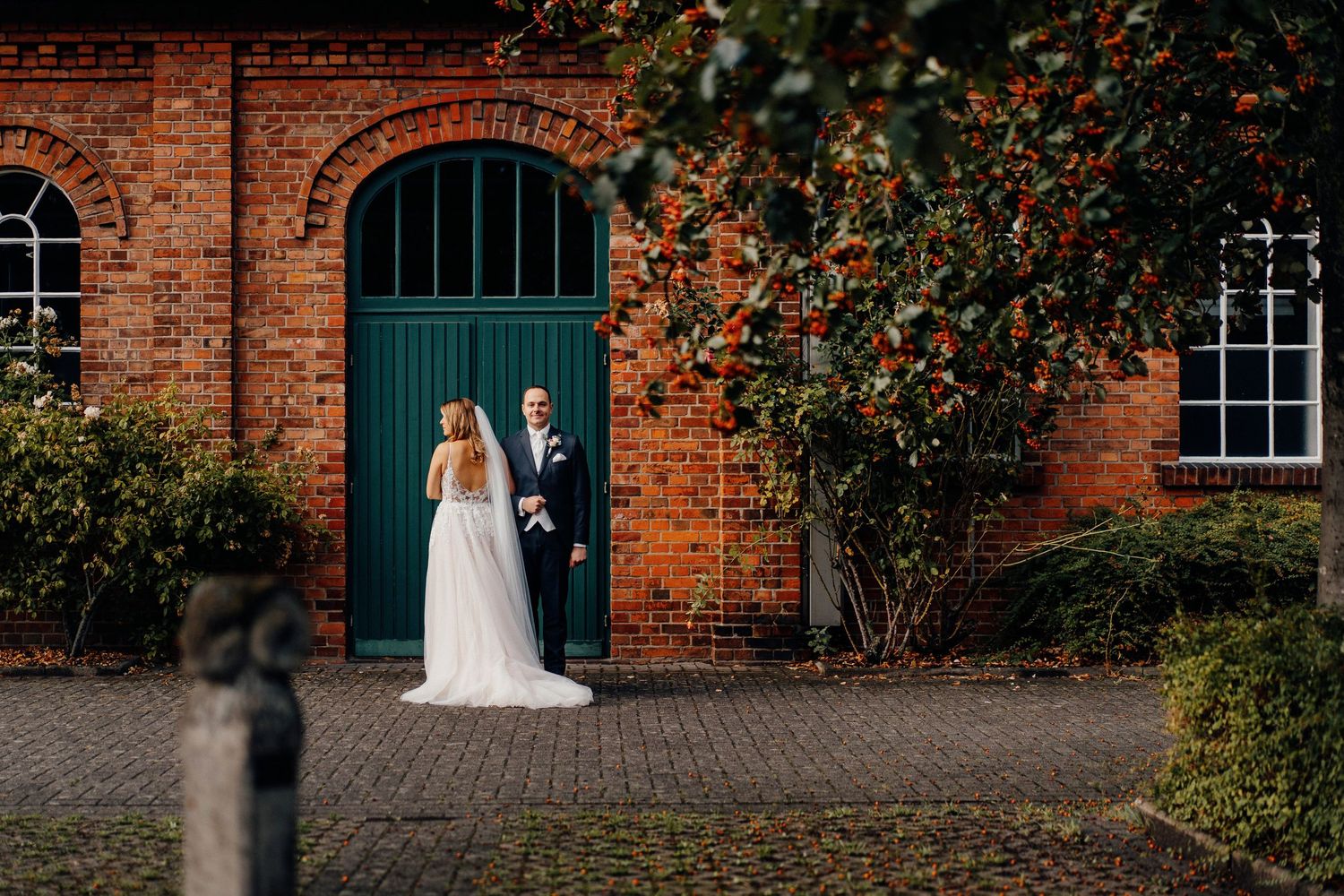 Wedding couple standing before a green door on a charming red brick building with lush greenery.