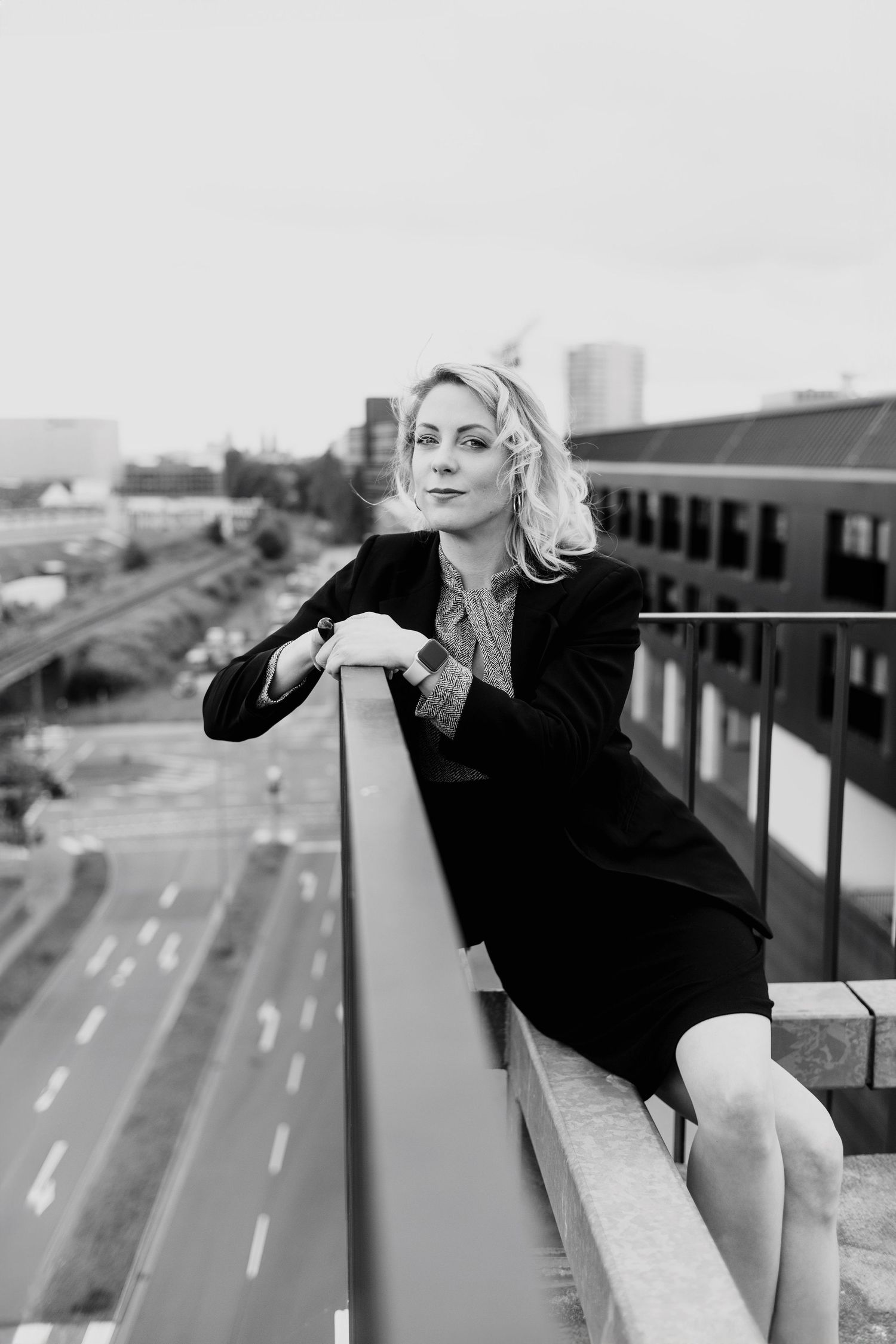 Elegant blonde woman in black suit leaning on rooftop railing with urban cityscape background.