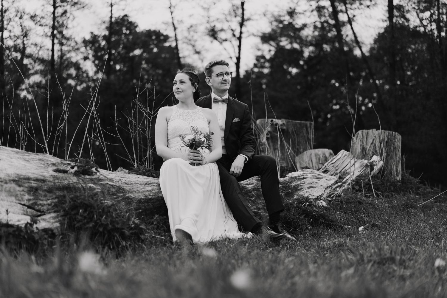 Black and white photo of bride and groom sitting together on a fallen log in a forest meadow.