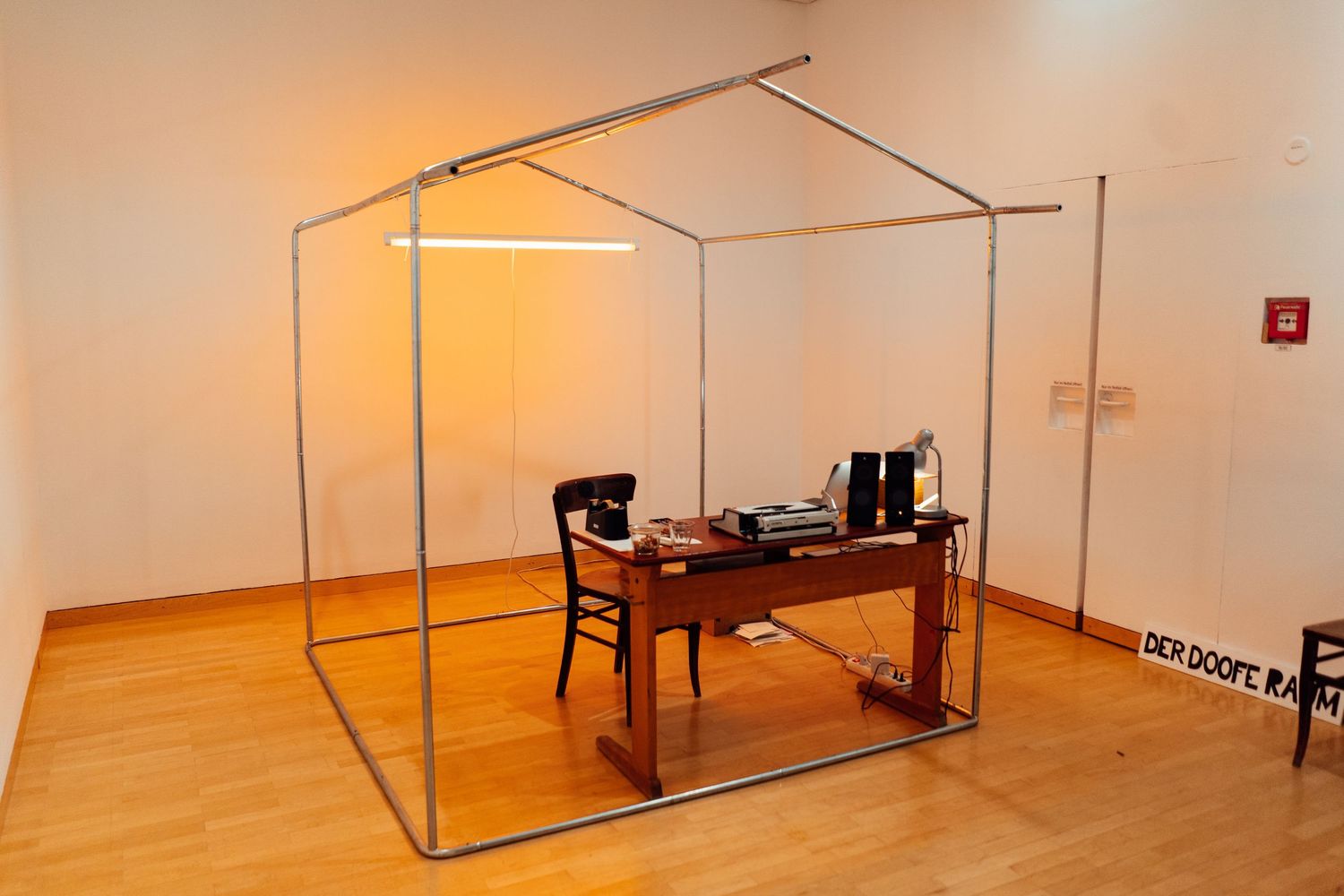 Art installation of a metal frame house structure surrounding a wooden desk in a gallery.
