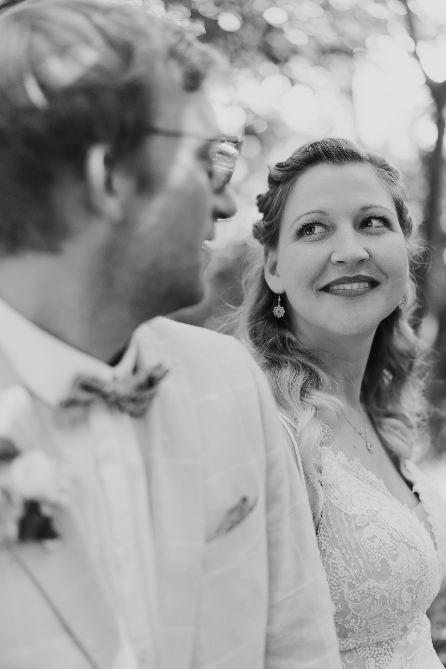 Black and white portrait of smiling bride gazing up at groom in light suit among trees.