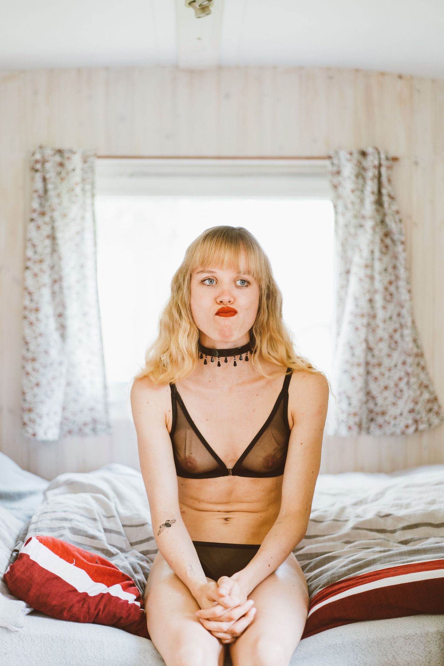 Blonde woman in sheer black lingerie sits on bed in retro camper trailer interior.
