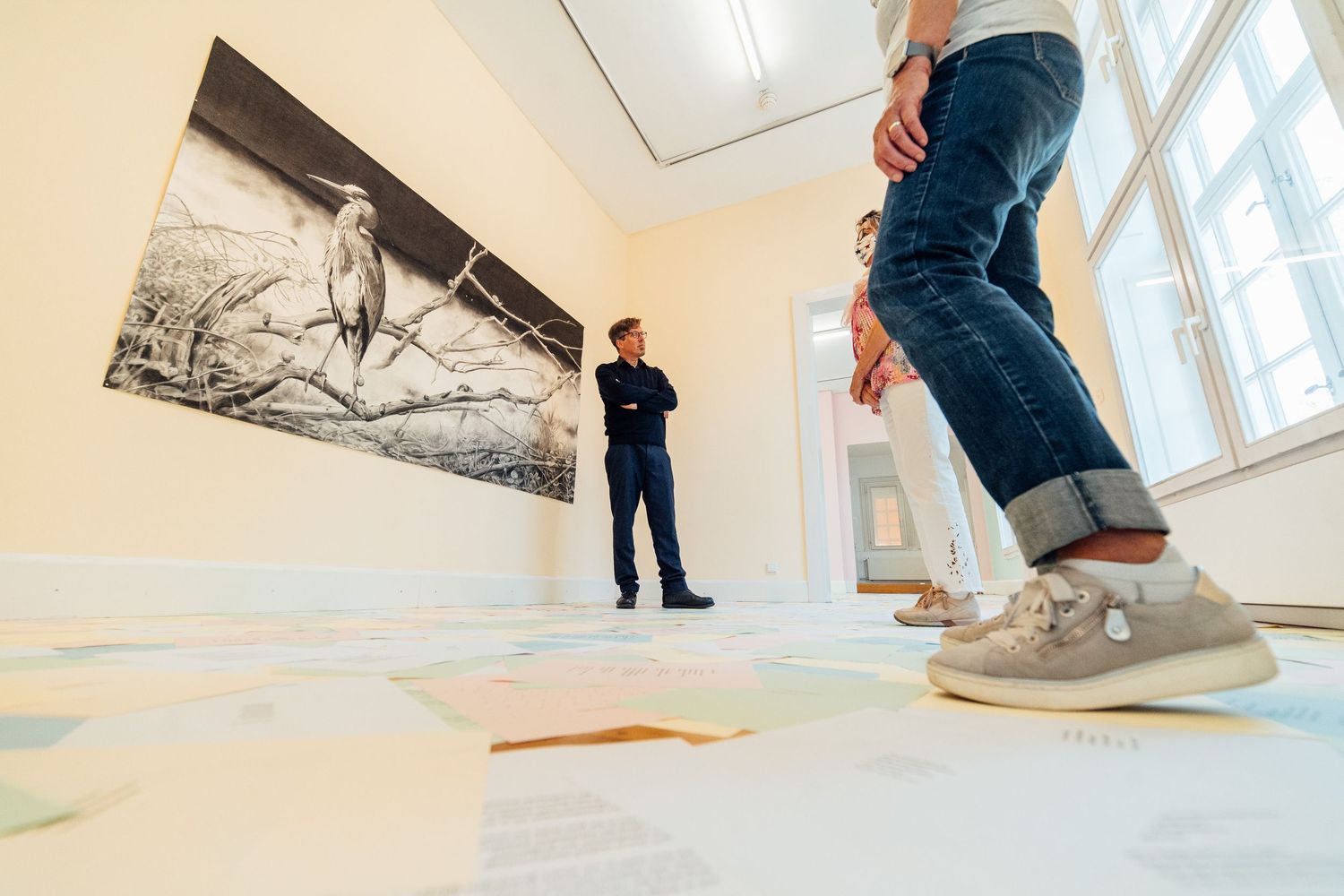Visitors viewing a large black and white photograph in a bright white gallery with colorful papers on the floor.