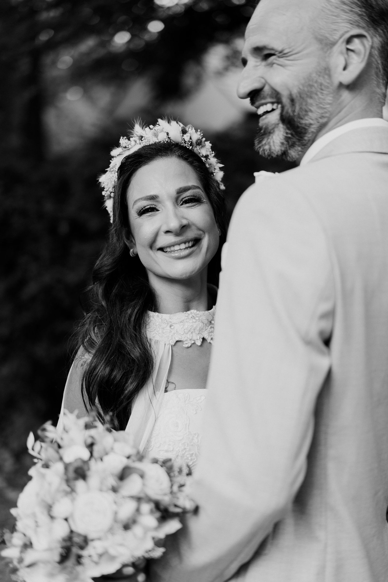 Black and white photo of a smiling bride with floral crown holding bouquet beside laughing groom in suit.