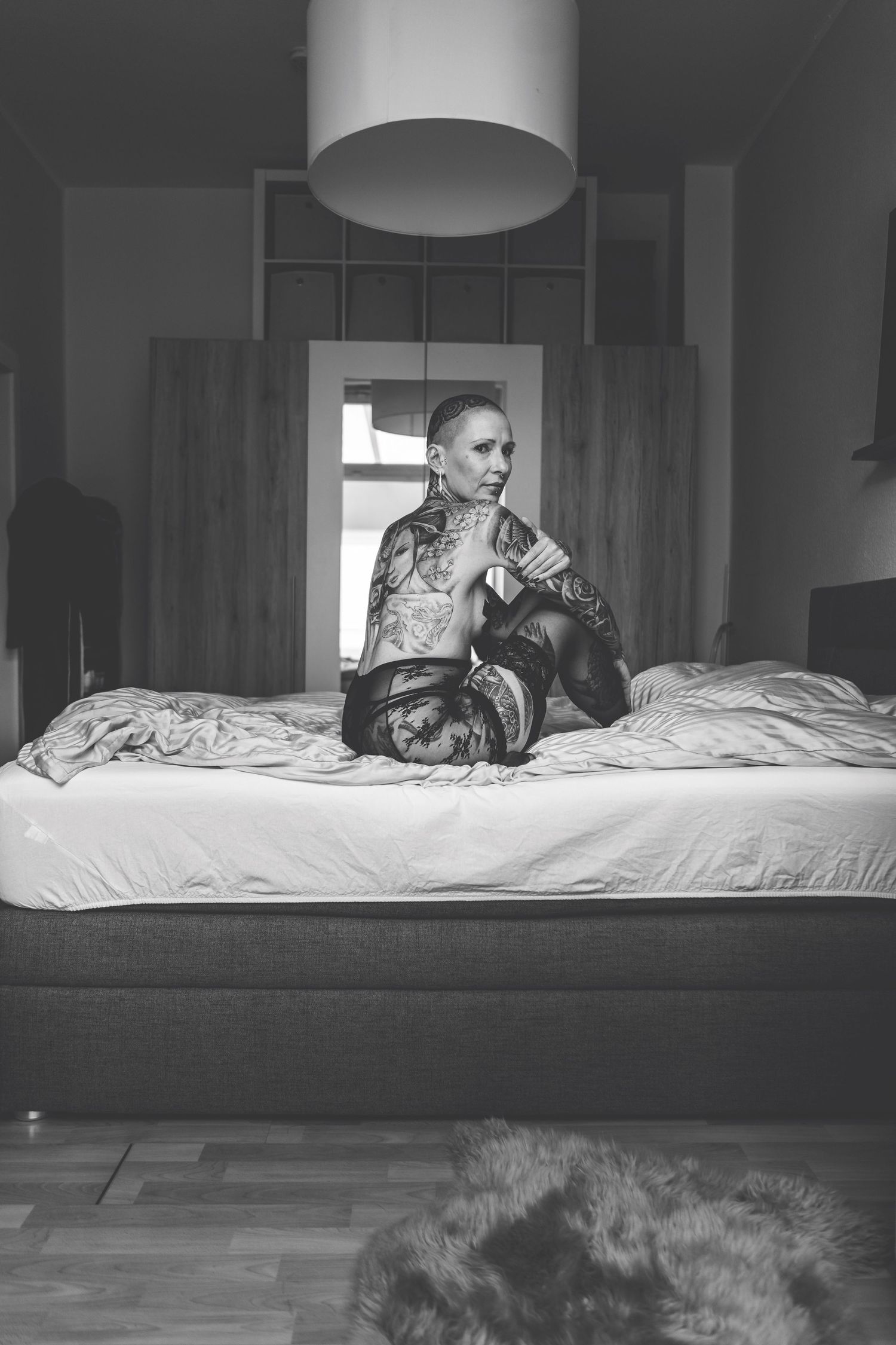 Black and white photo of tattooed bald woman kneeling on bed in lace lingerie.