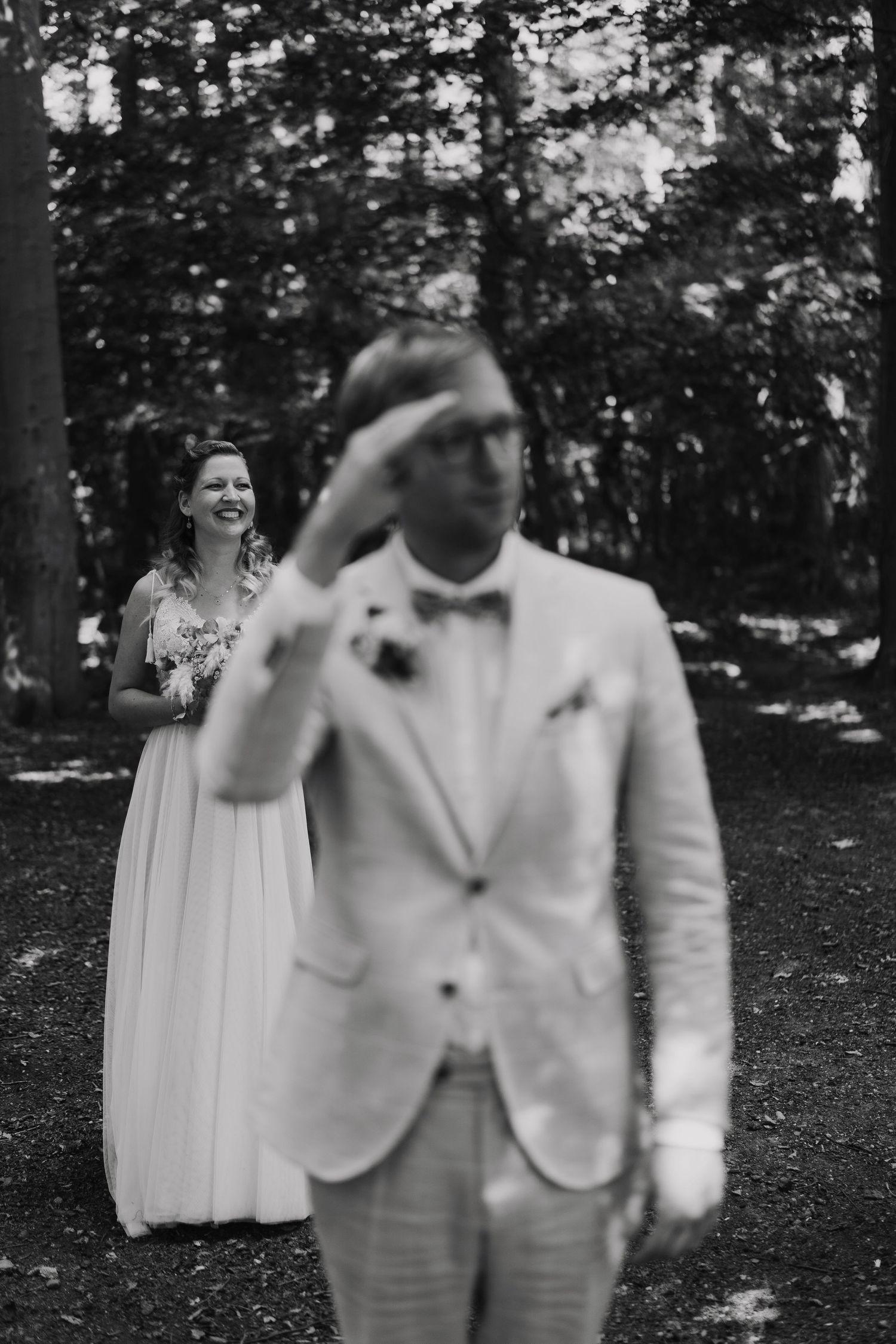 Black and white wedding photo of smiling bride approaching groom from behind in a forest setting.