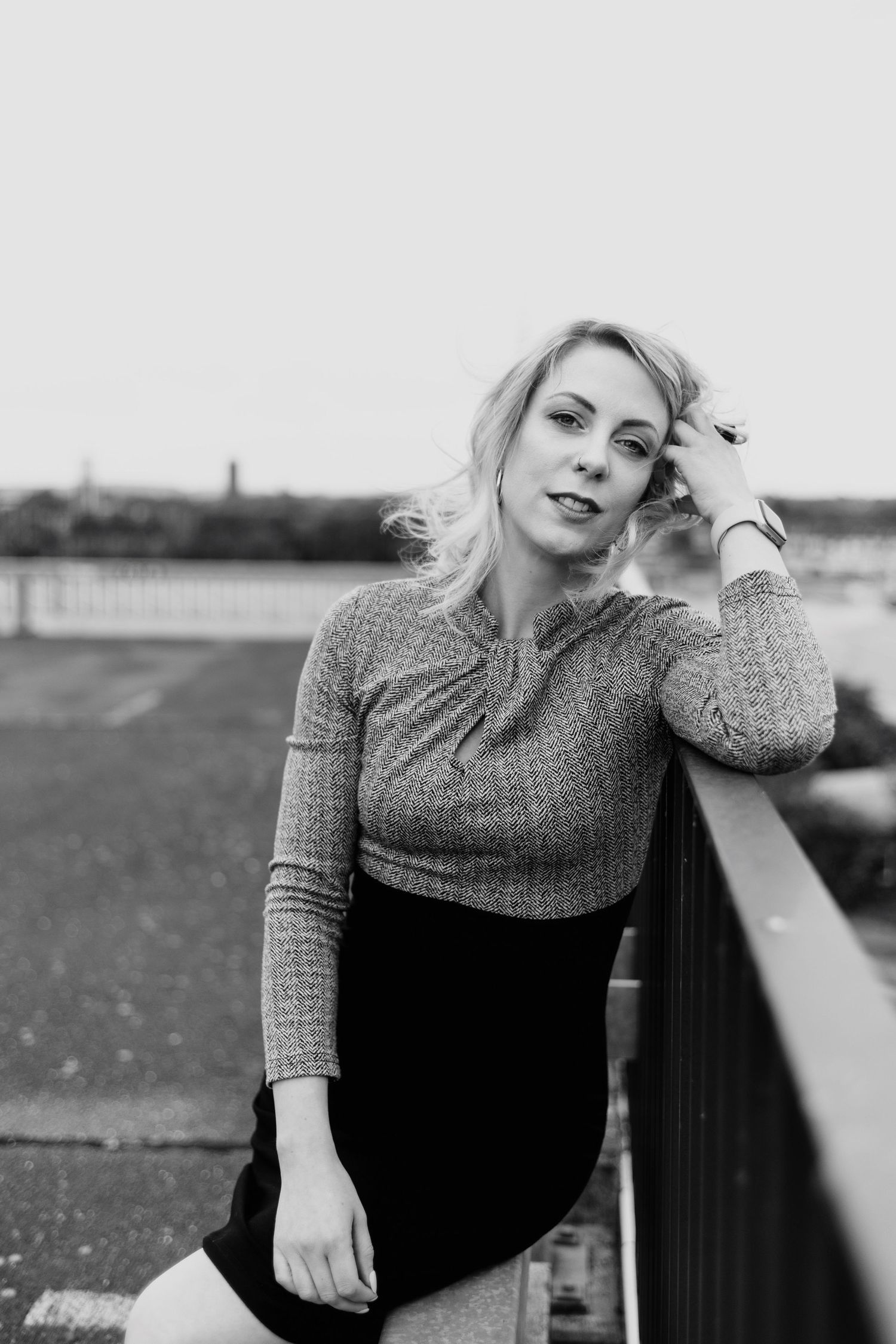 Black and white portrait of blonde woman in sweater and skirt leaning on railing outdoors.