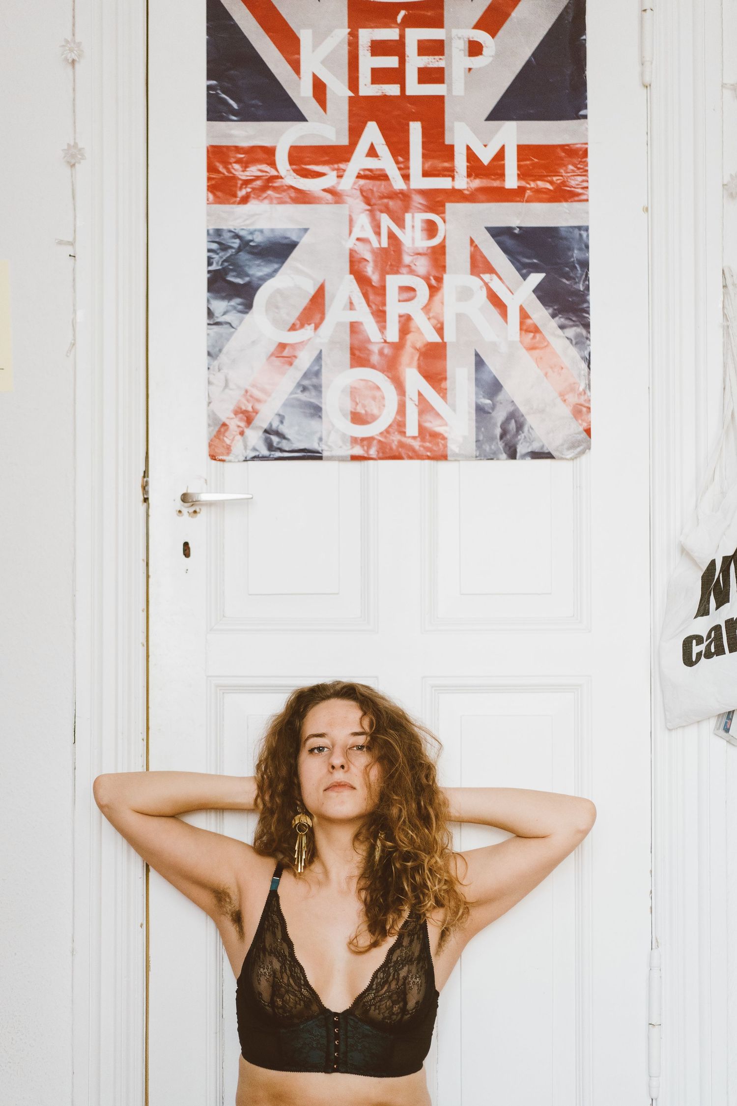 Young woman in black lace bralette standing against white door with Keep Calm and Carry On poster.