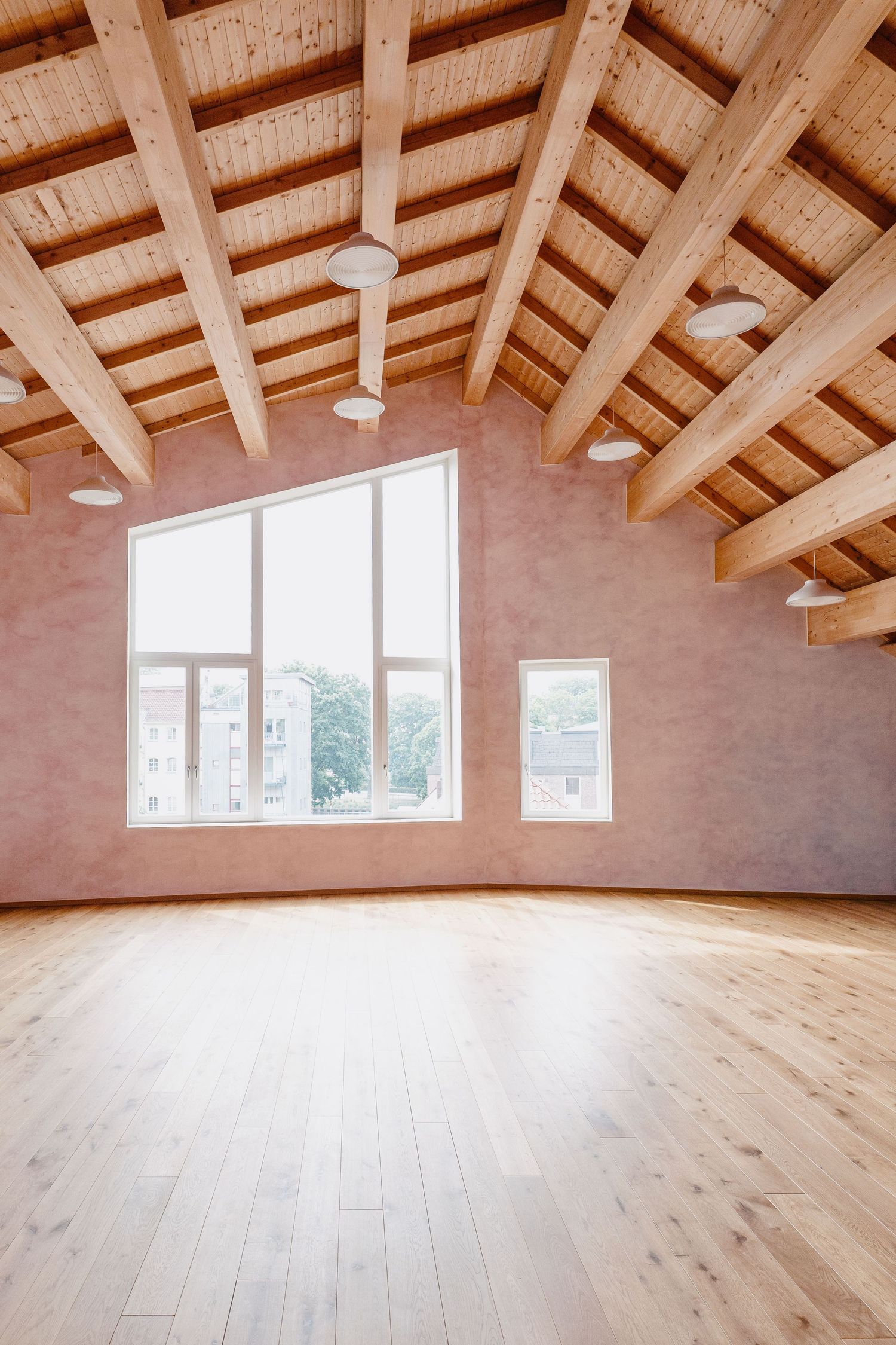 Empty room with exposed wooden beam ceiling, large windows, and light hardwood floors.