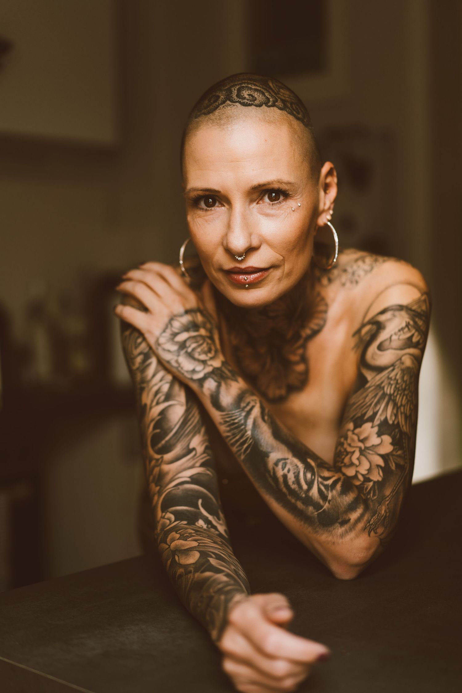 Portrait of tattooed bald woman with nose ring leaning on dark surface in warm light.