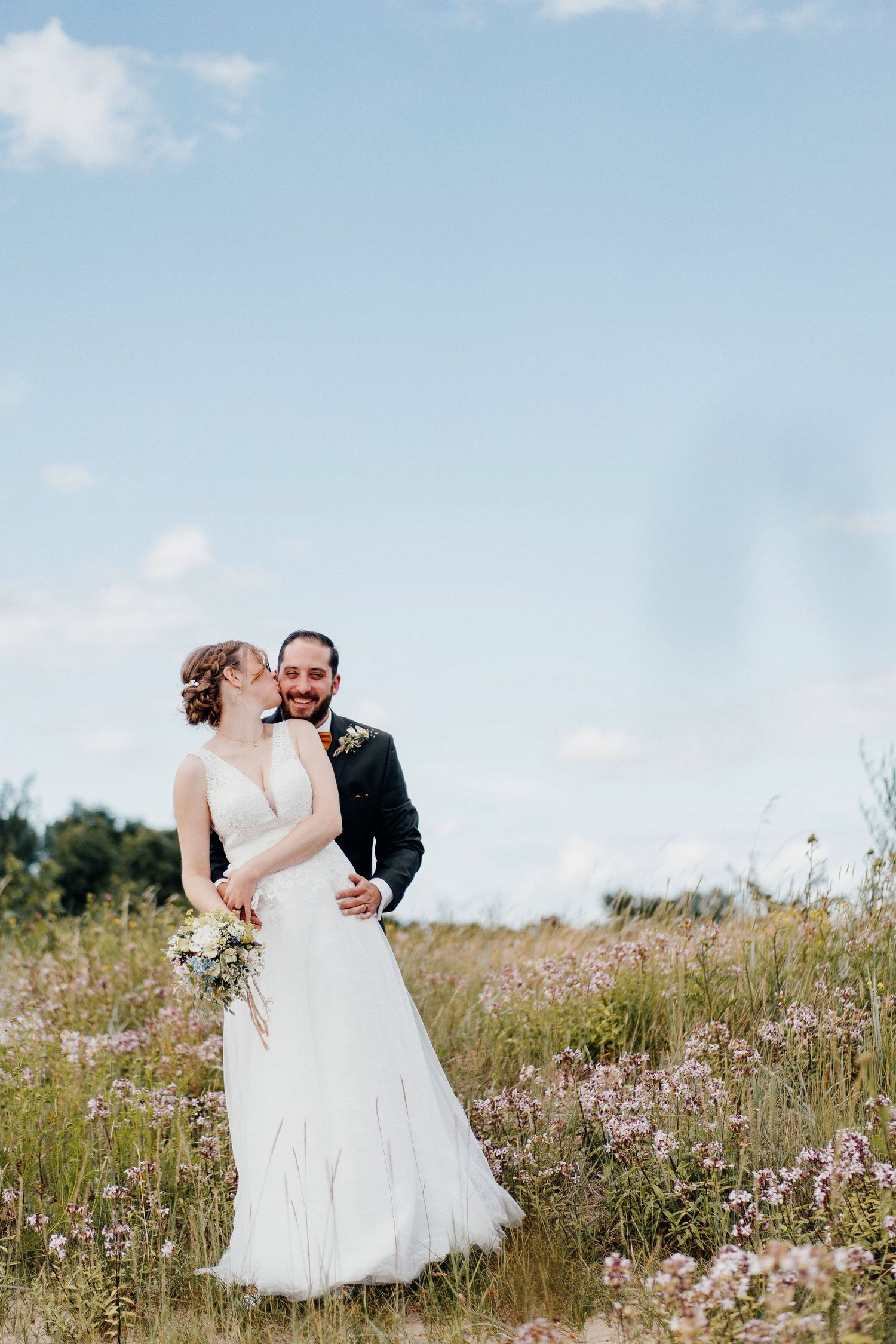 Bride and groom embracing in a blooming wildflower field under a bright blue summer sky.