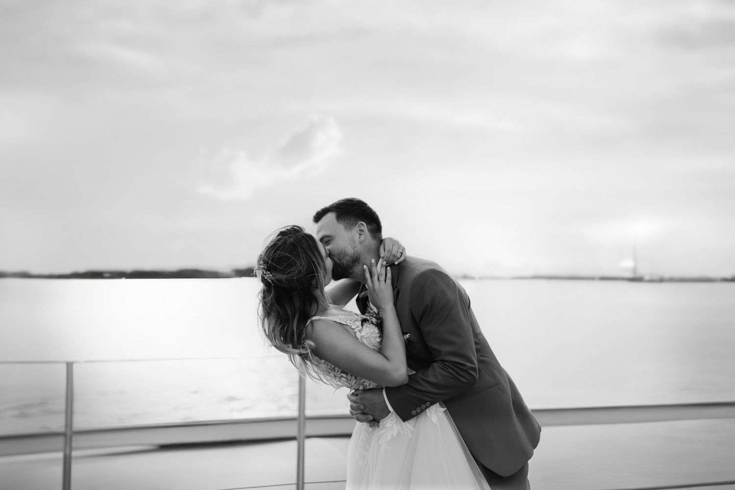 Black and white romantic photo of newlyweds sharing a kiss by the waterfront at sunset.