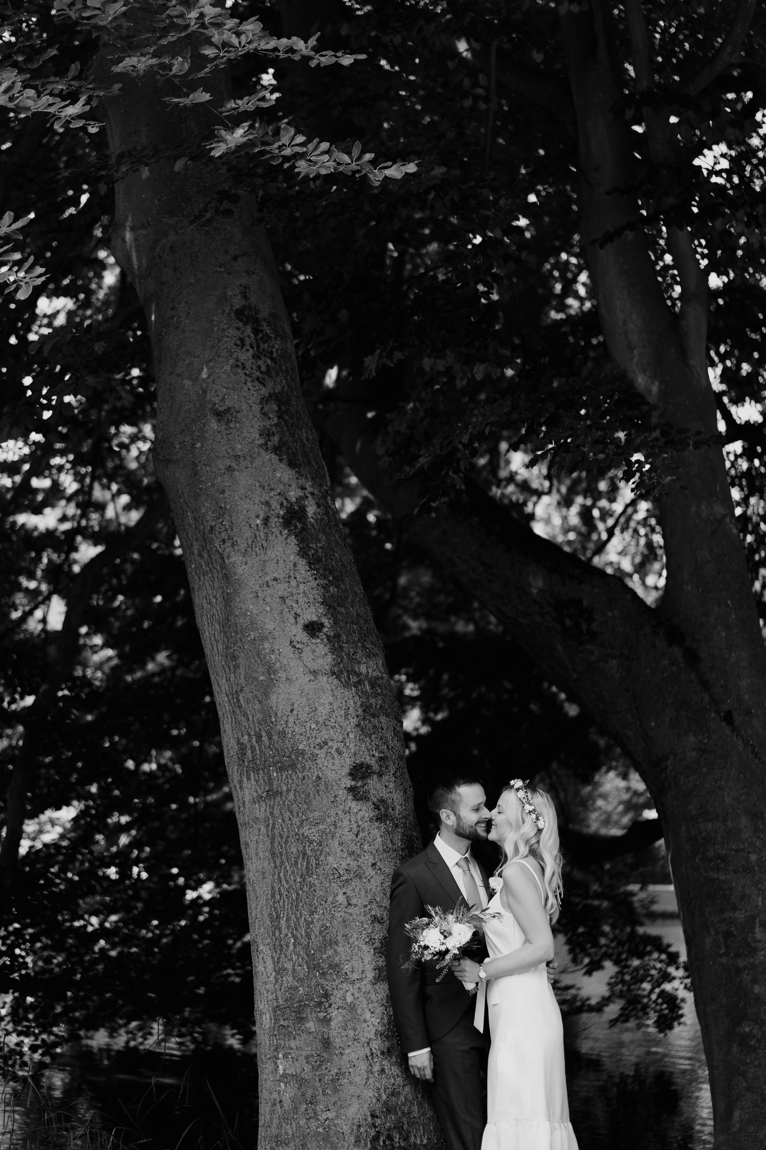 Black and white photo of bride and groom sharing intimate moment beneath large tree trunk.