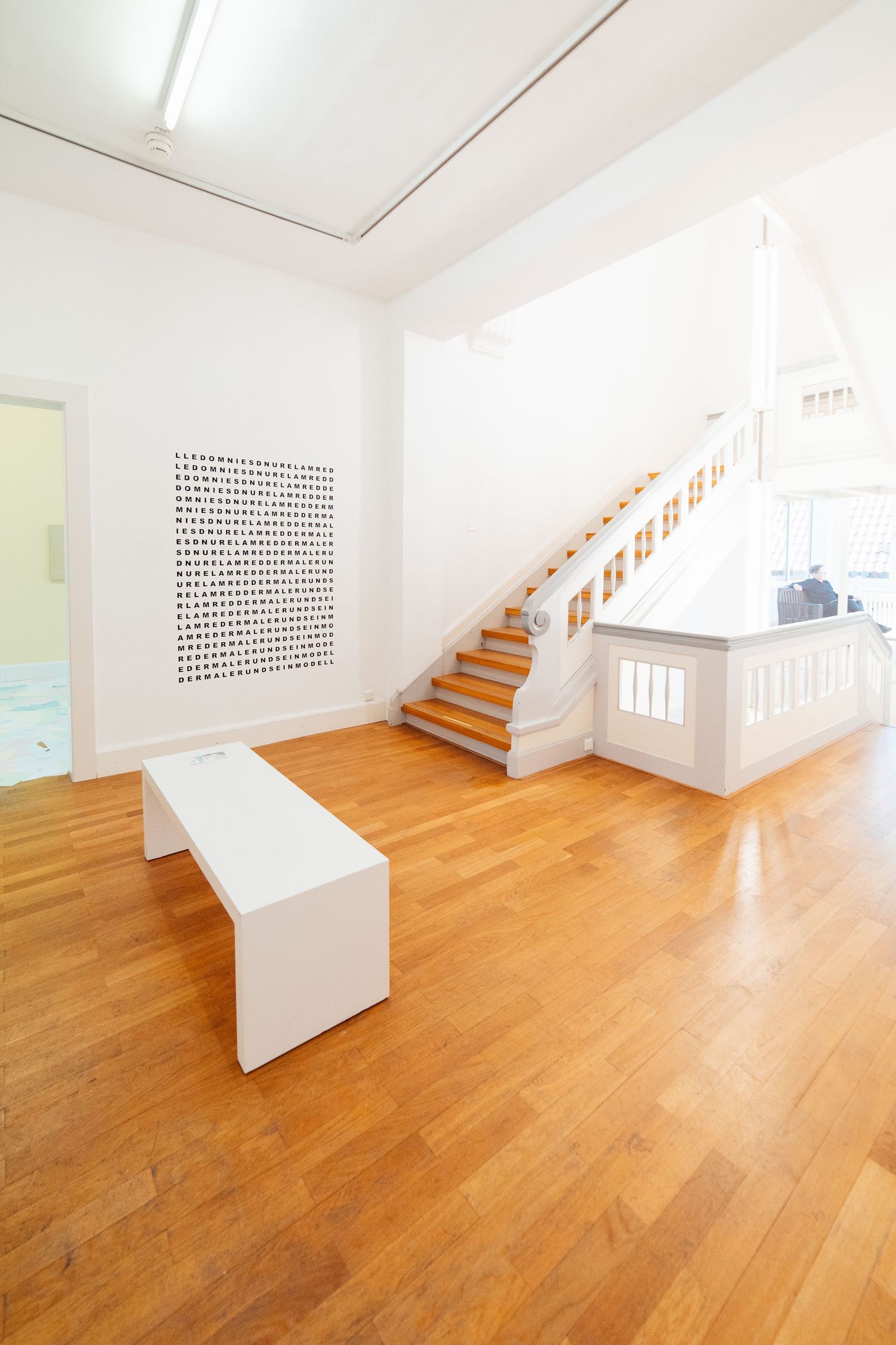 Minimalist white gallery space with wooden staircase, parquet floor, and abstract dot artwork on the wall.