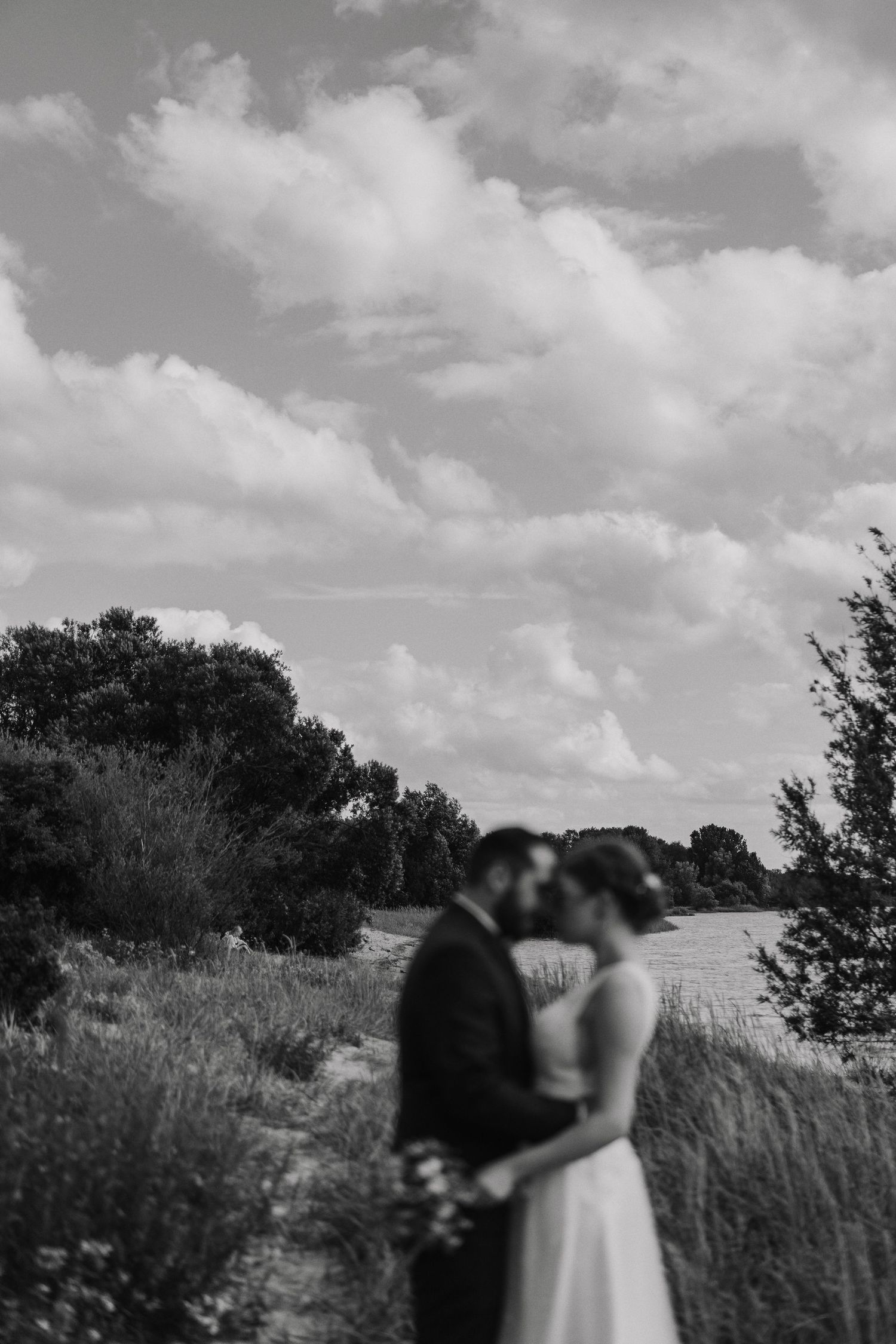 Black and white photo of bride and groom kissing in a wildflower field under dramatic cloudy sky.