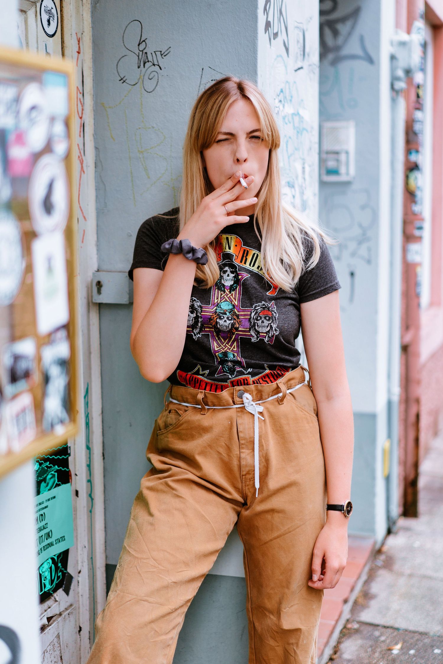 Blonde woman smoking cigarette leaning on graffiti wall outdoors.