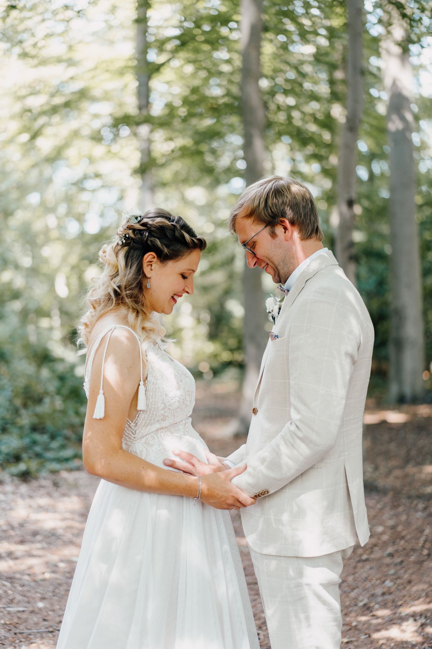 Smiling bride and groom in forest holding hands, bride in white lace dress, groom in cream suit.
