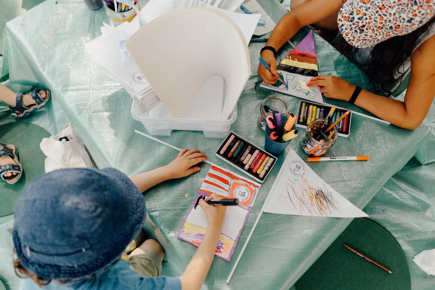 Children drawing and coloring with pastels and markers at a table covered with a light blue tablecloth.