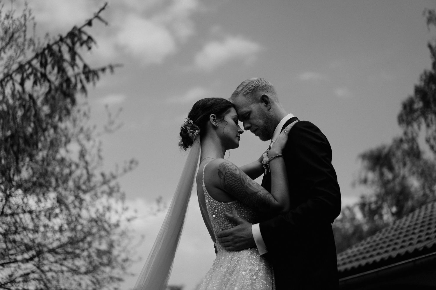 Bride and groom share intimate moment forehead to forehead under cloudy sky in black and white wedding photo.