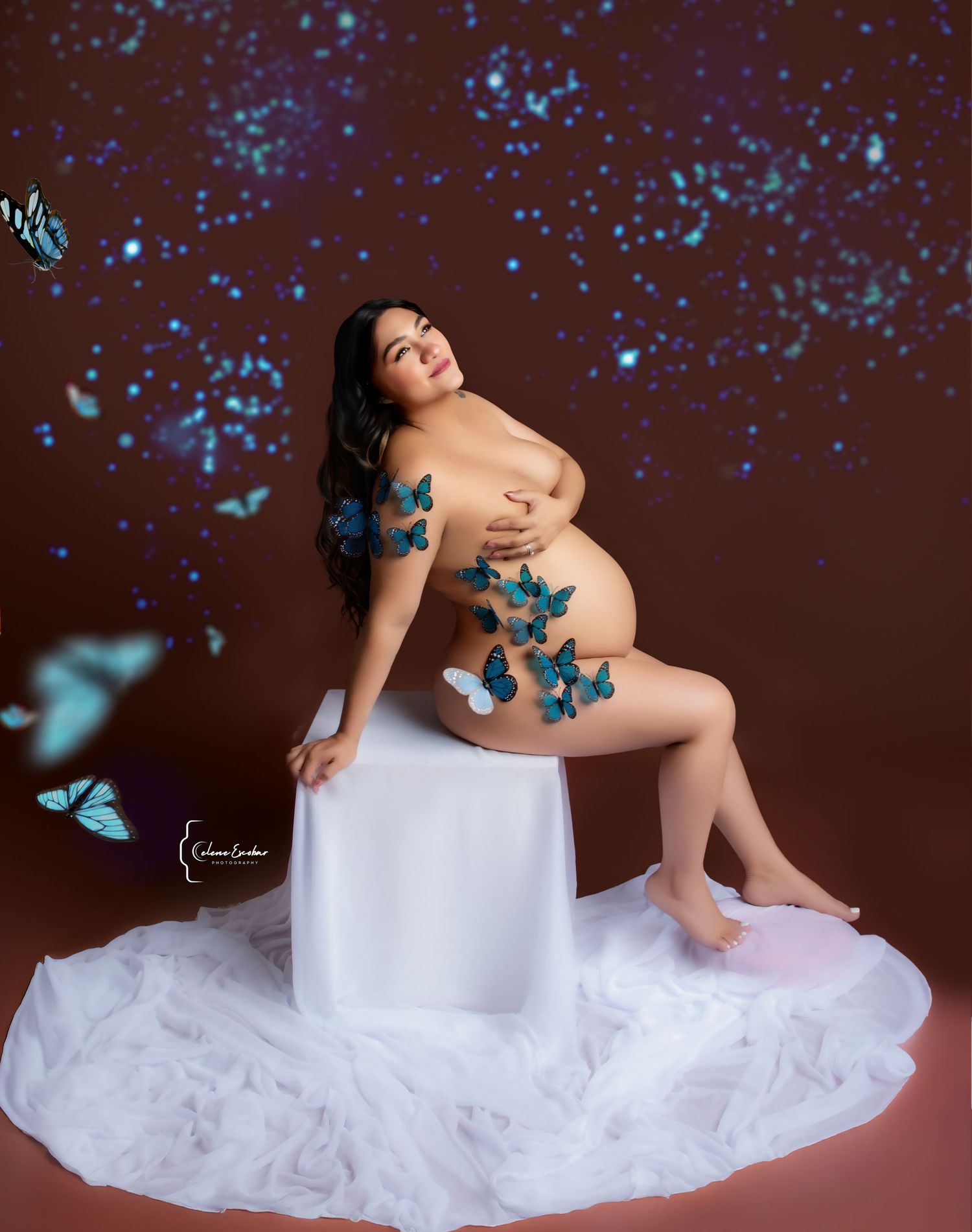 Artistic maternity photo with a pregnant woman adorned with blue butterflies on dark background.