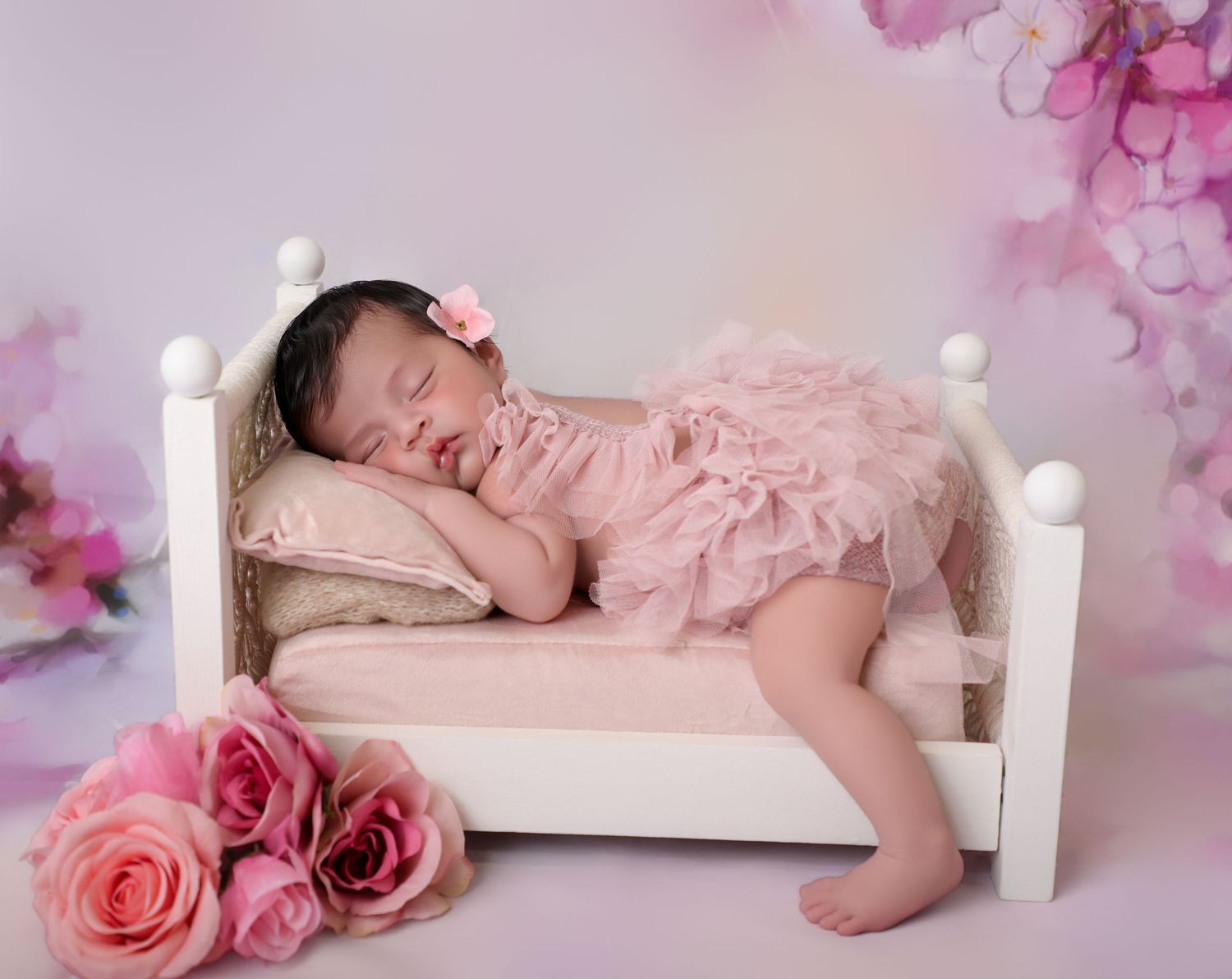 Sleeping newborn baby in pink tutu dress rests on a tiny white bed surrounded by pink roses in a studio.