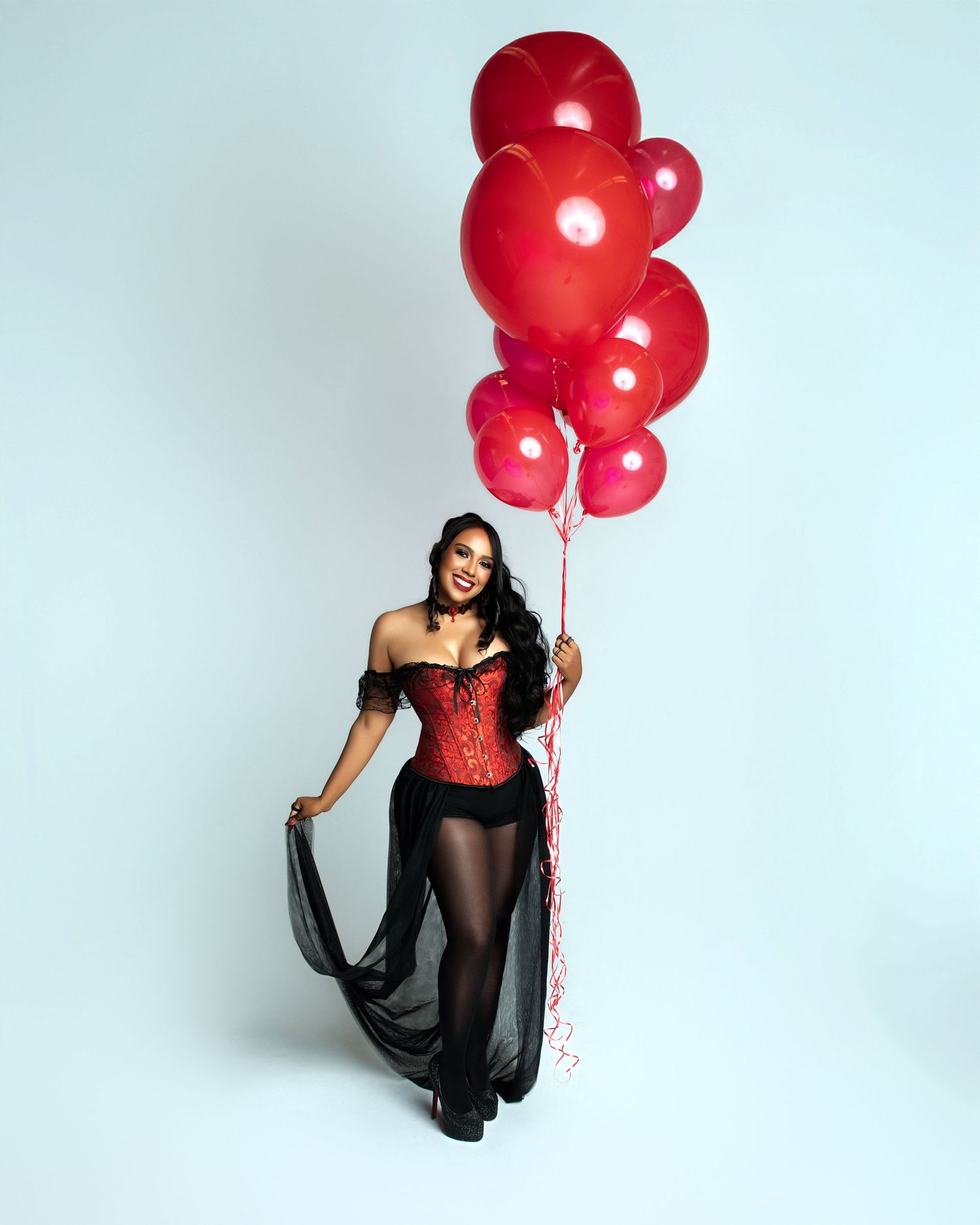 Woman in red corset and black sheer skirt holds red balloons, smiling against light blue background.