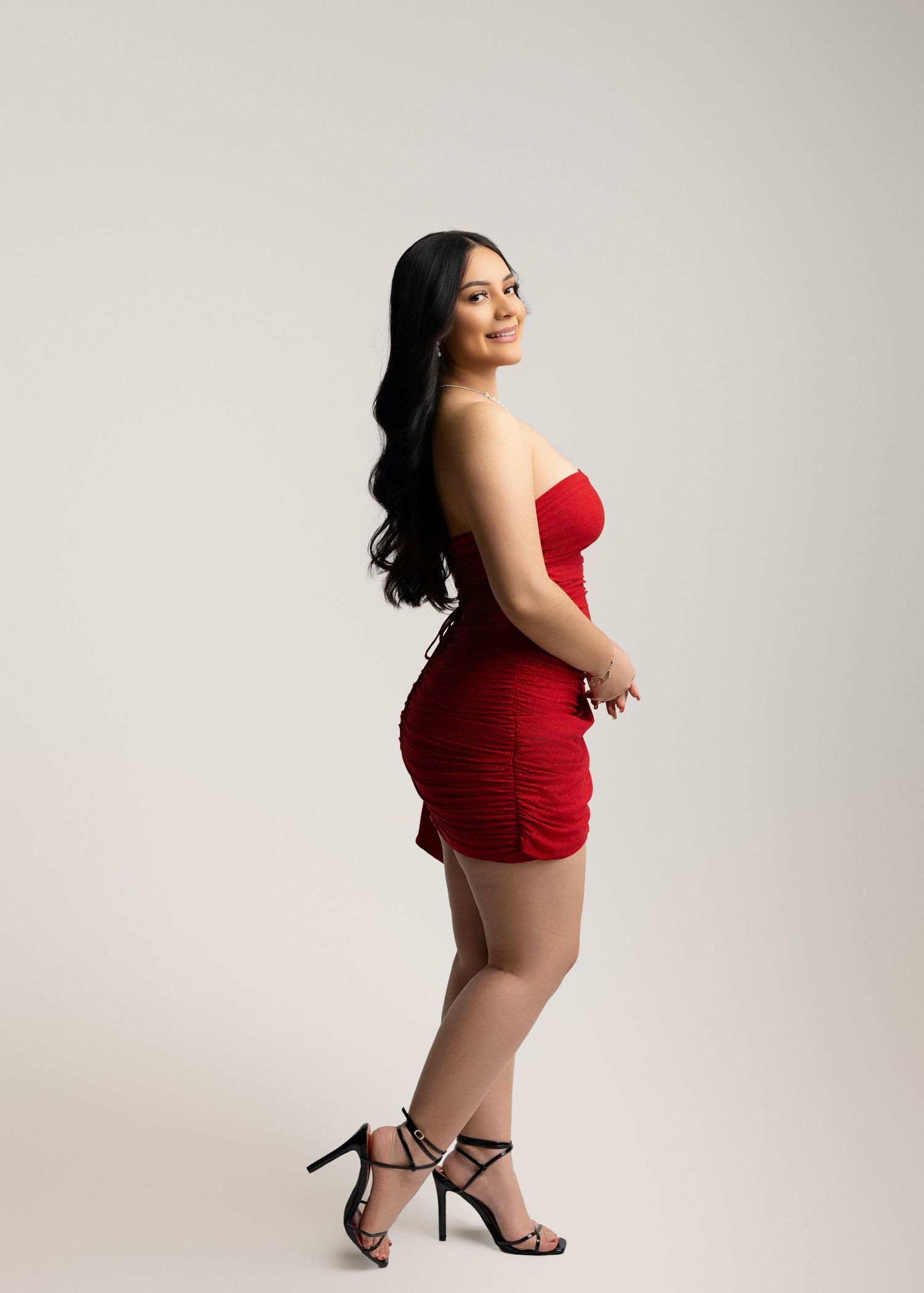 Woman in red strapless ruched mini dress and black strappy heels posing sideways against a white background.