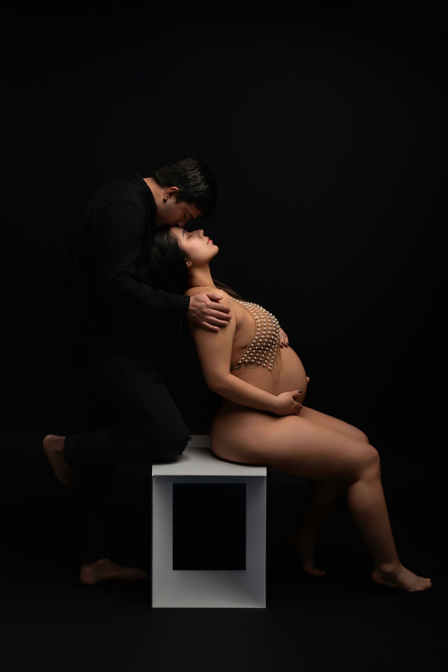 Couple sharing intimate maternity photo, pregnant woman in pearl bodysuit seated on white cube, partner in black suit kissing her forehead.