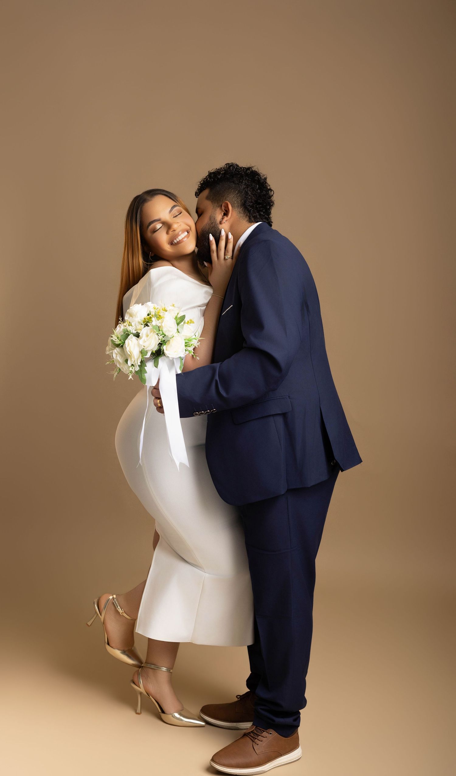A couple in wedding attire embraces, bride in white dress holding bouquet, groom in navy suit, against beige background.