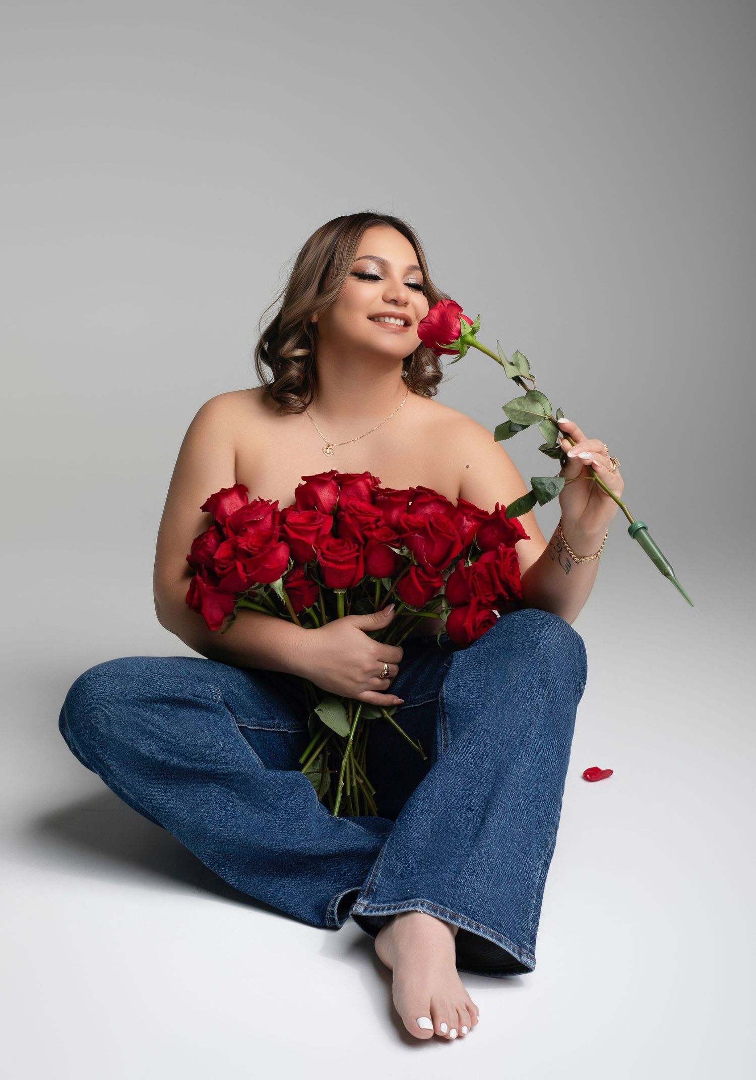 Woman sitting on floor holding red roses bouquet, wearing jeans, smiling and smelling a single rose on grey background.