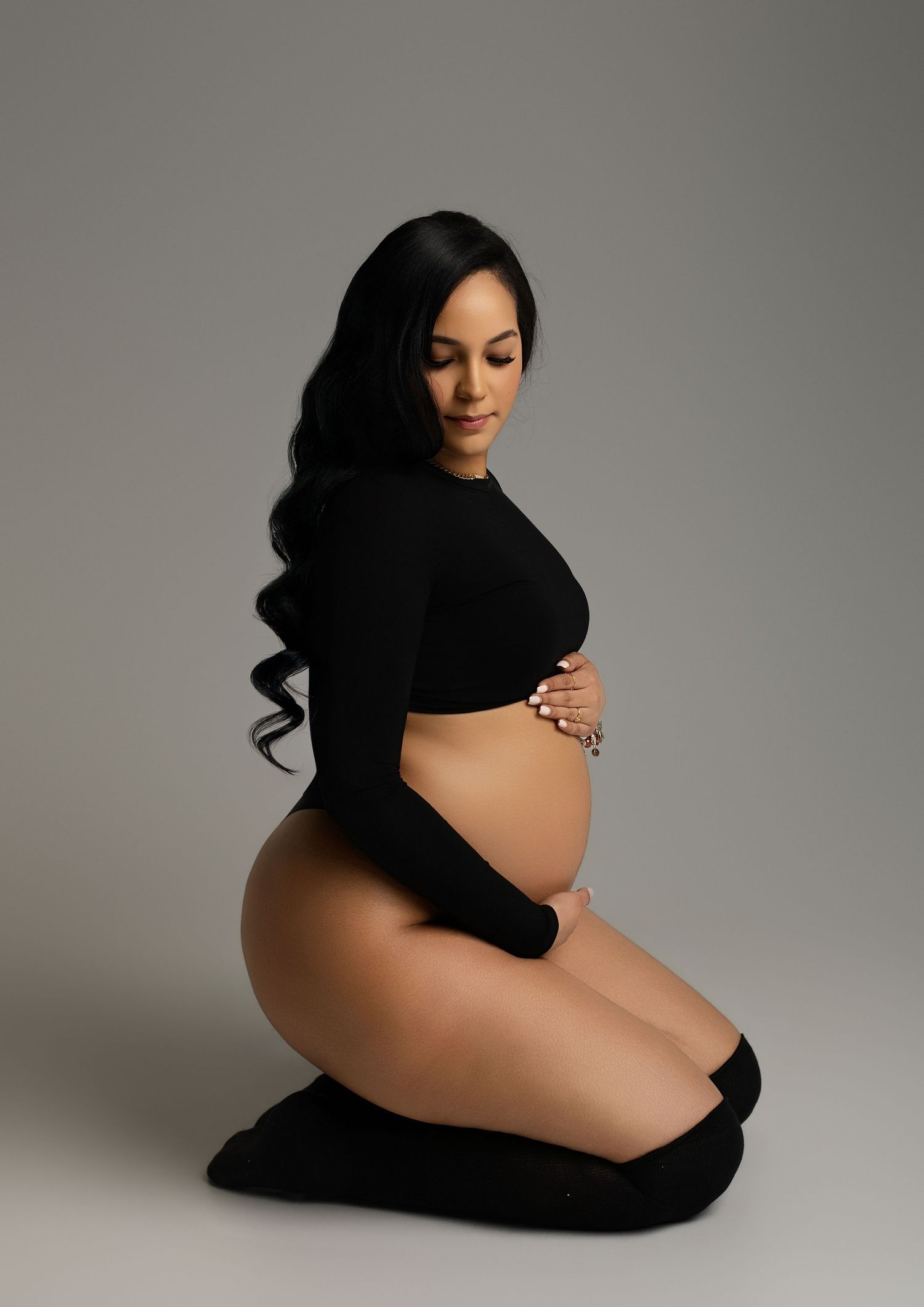 Pregnant woman in black crop top kneeling gracefully, cradling her baby bump during a professional maternity photo session.