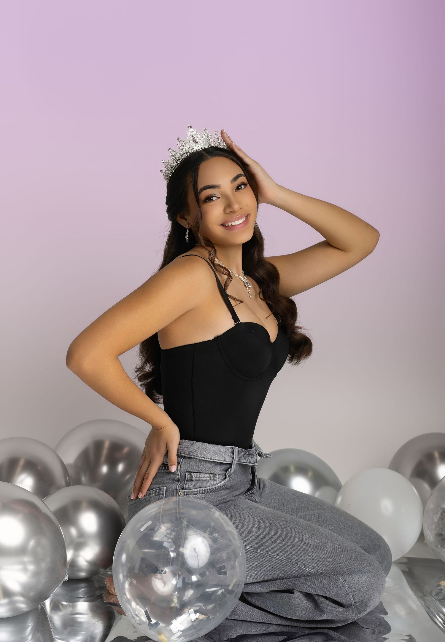 Young woman wearing a crystal crown and black bodysuit sits among silver balloons on a pink gradient background.