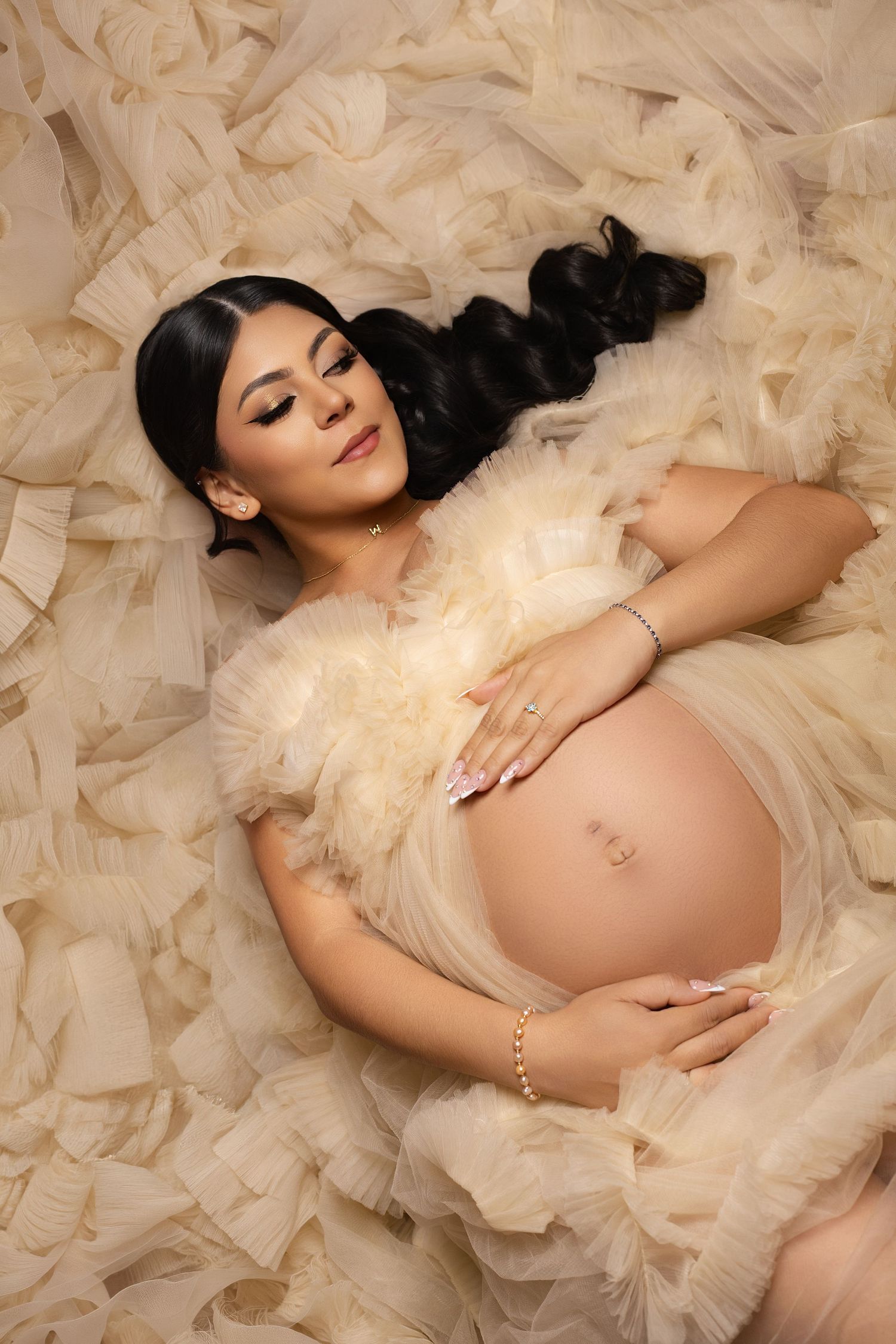 Pregnant woman in cream tulle gown lying down, cradling baby bump with manicured hands and elegant bracelet.