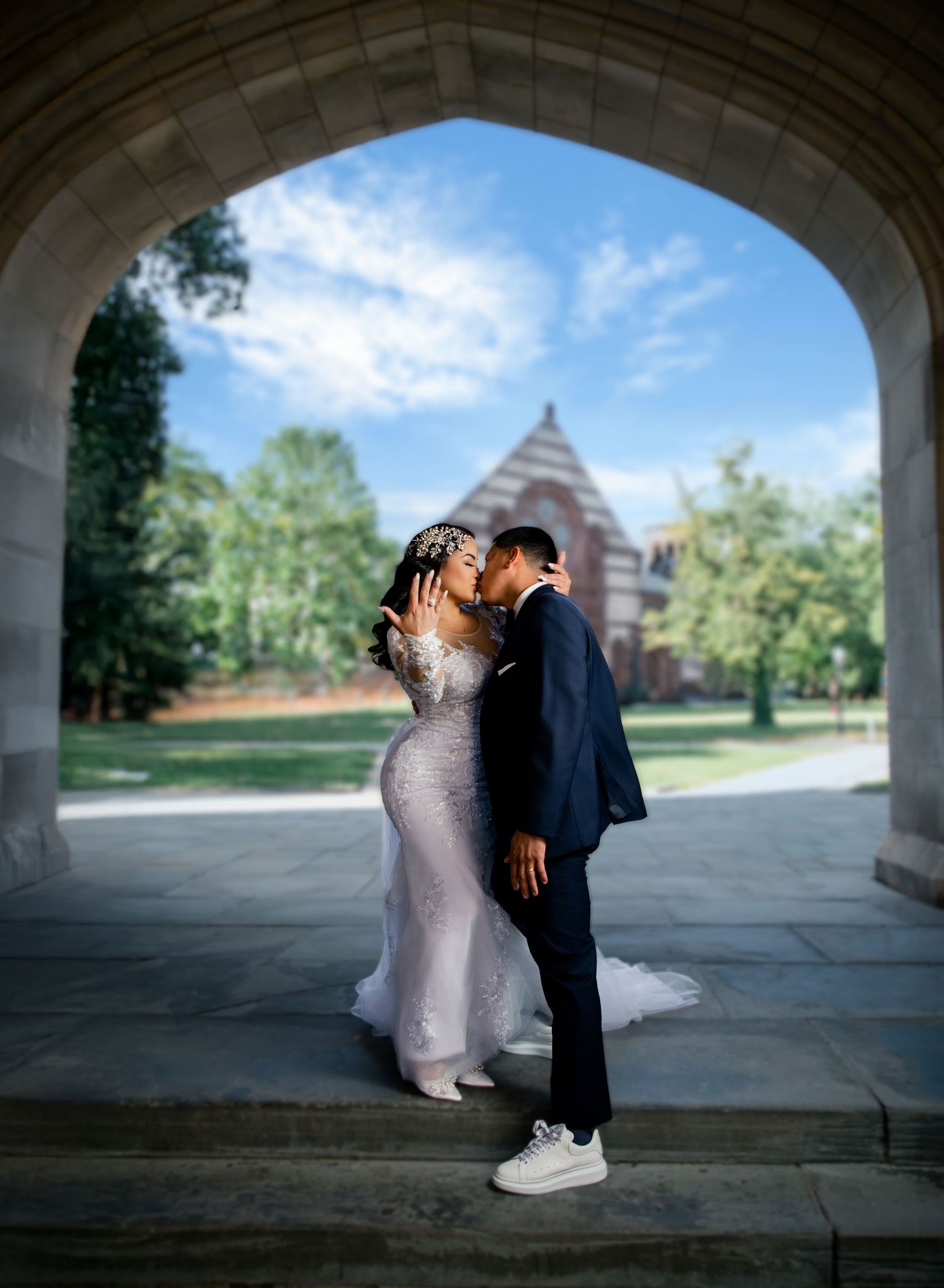 A couple shares a romantic kiss in wedding attire under a stone arch, with lush greenery and a historic building backdrop.
