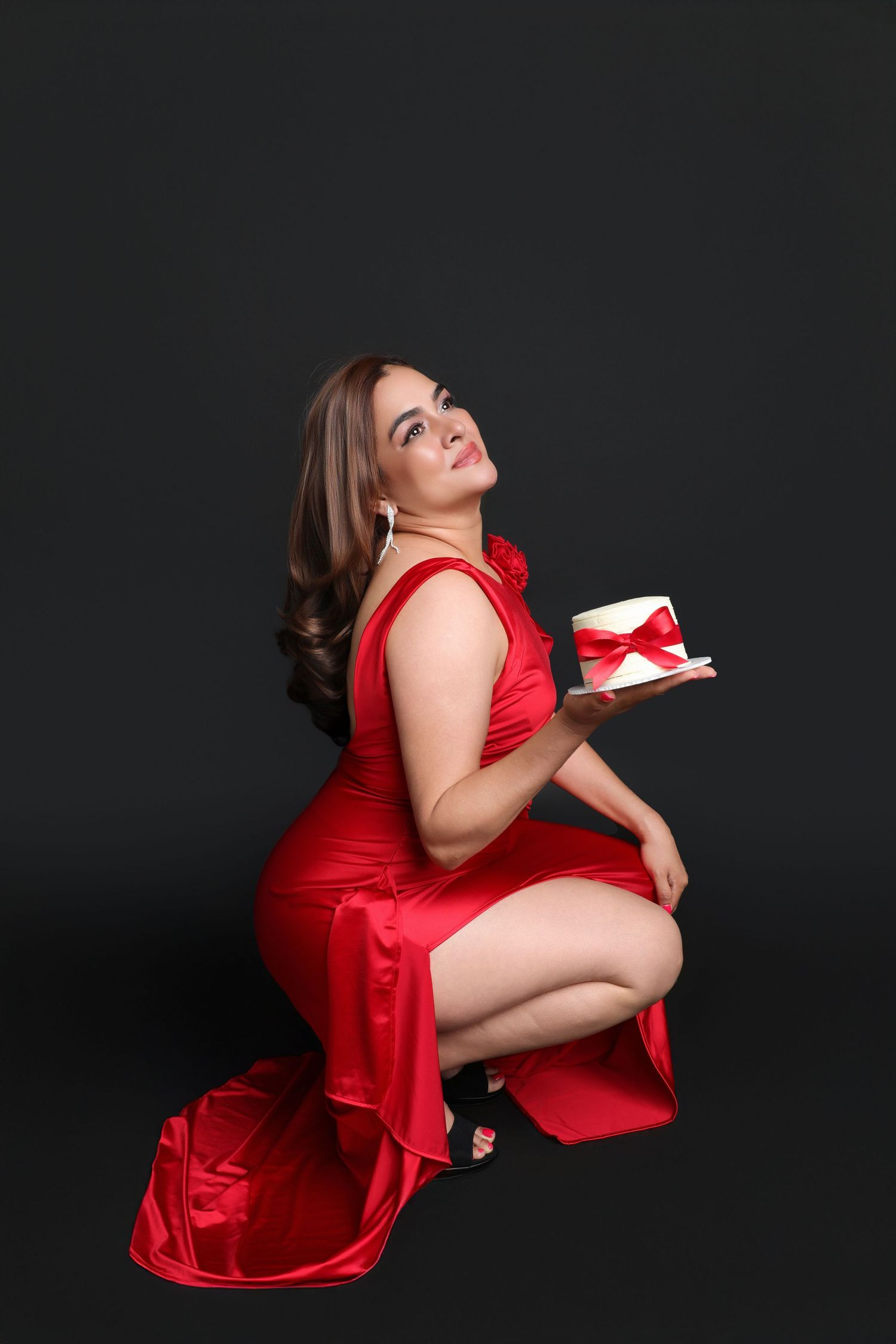 Woman in a red satin gown posing with a small white gift cake on a dark background.