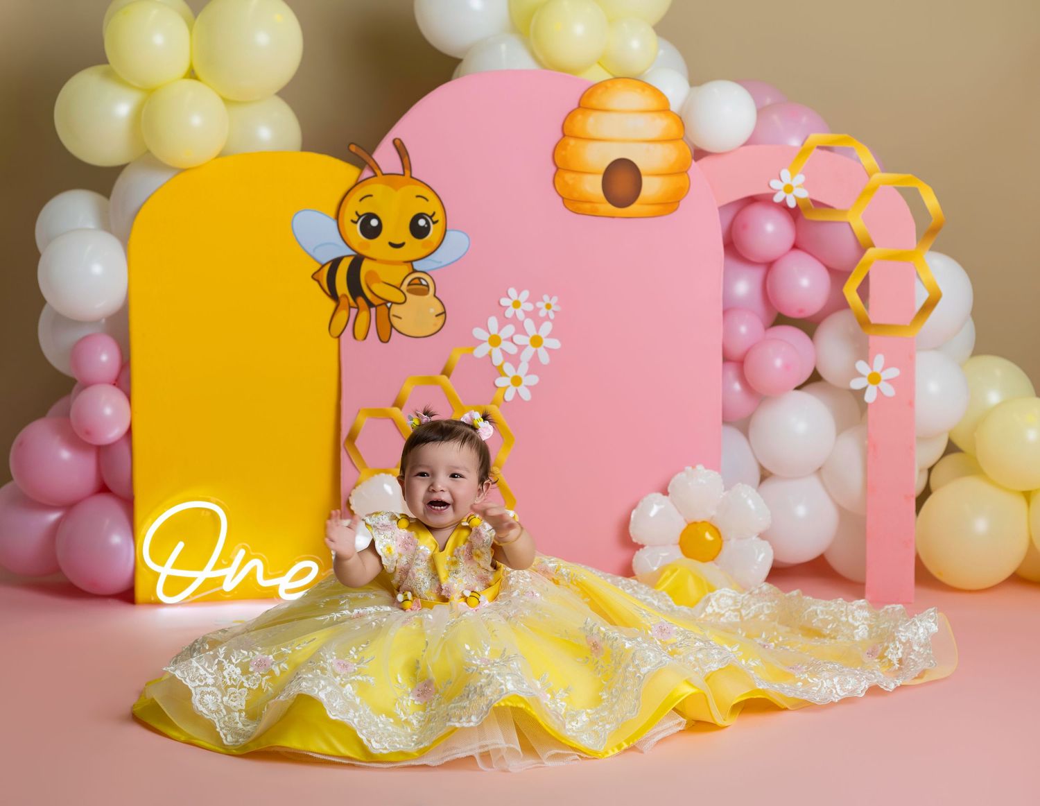Baby girl in yellow dress at bee-themed first birthday party with pink backdrop, honeycomb decor, and balloon garland.