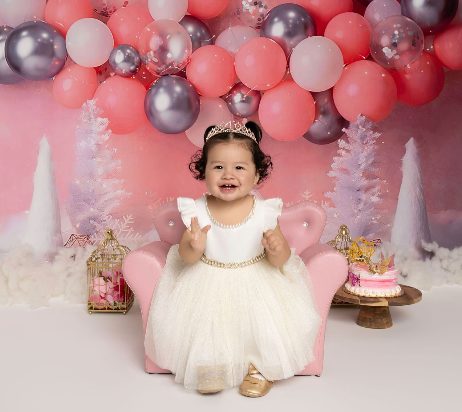 Smiling baby girl in white dress and tiara sits on pink chair surrounded by balloons and birthday cake decorations.