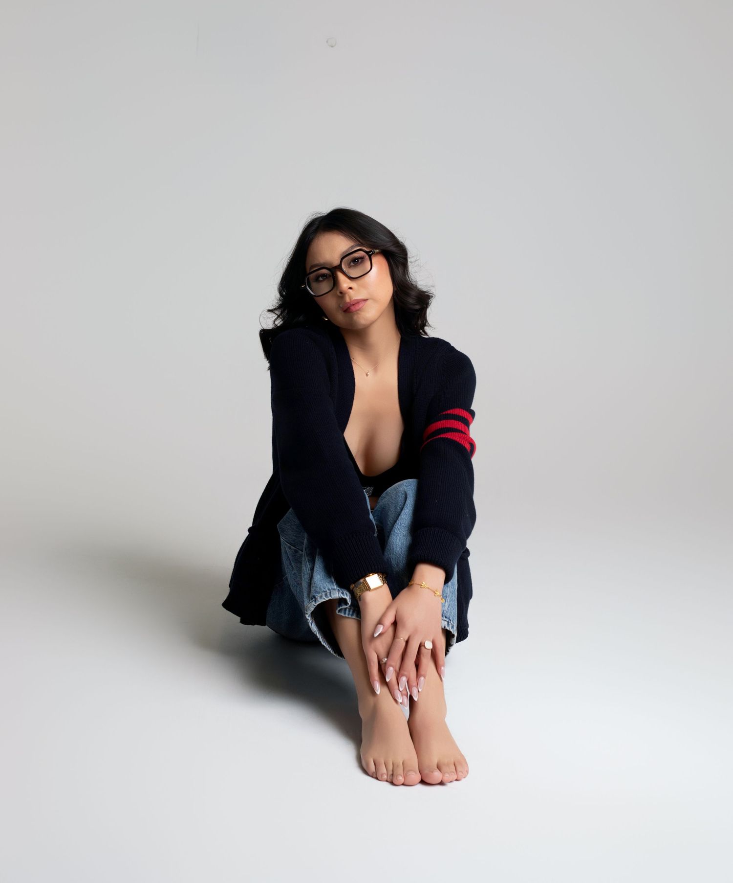 Young woman in navy cardigan with red stripes and jeans sits barefoot on white floor, wearing glasses.