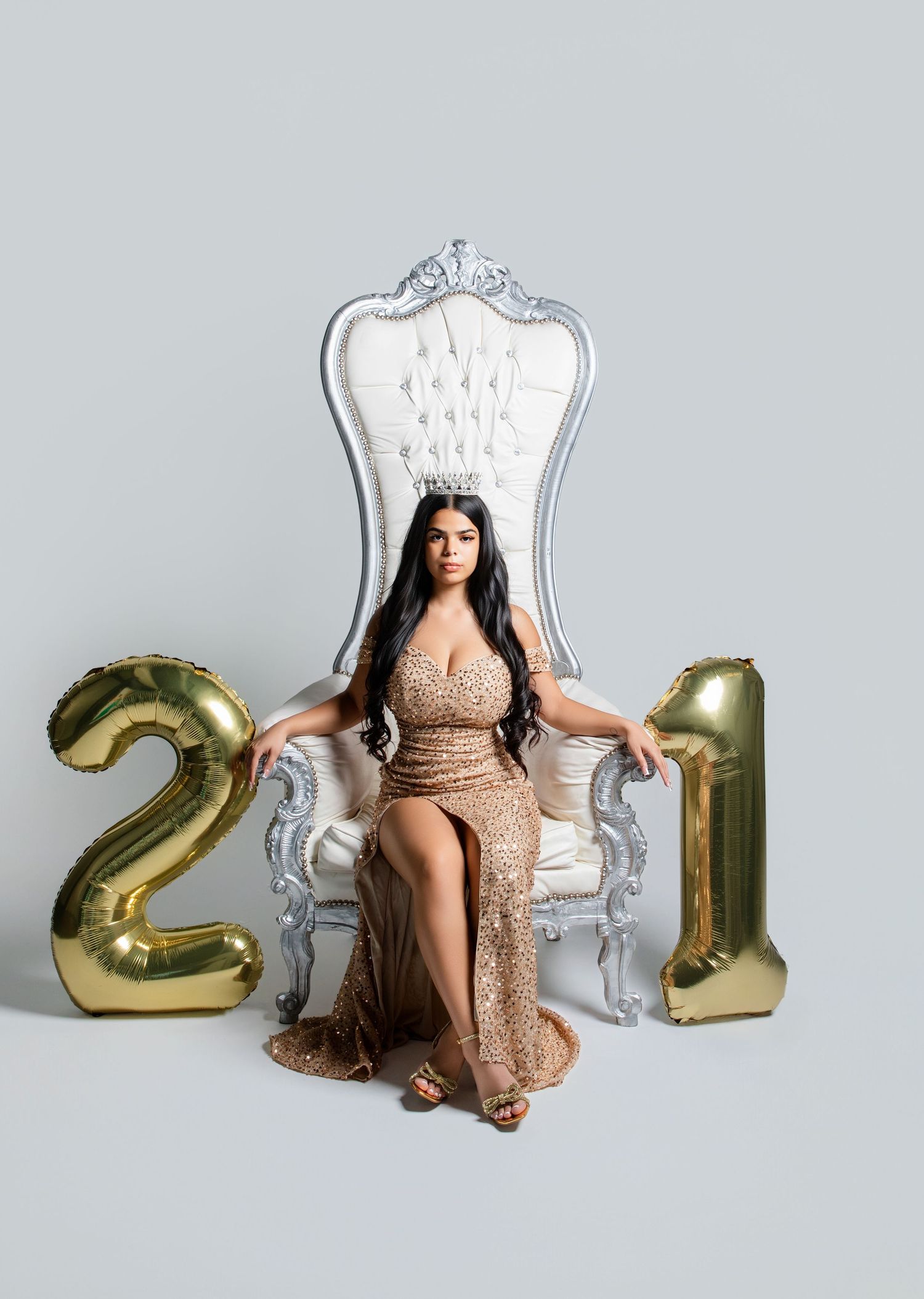 Woman in gold sequin gown seated on ornate throne chair with gold number 21 balloons for birthday celebration.