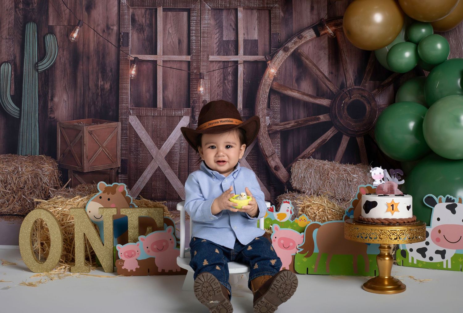Baby boy in cowboy hat sits among farm-themed first birthday decorations with hay bales, cow cake, and balloon backdrop.