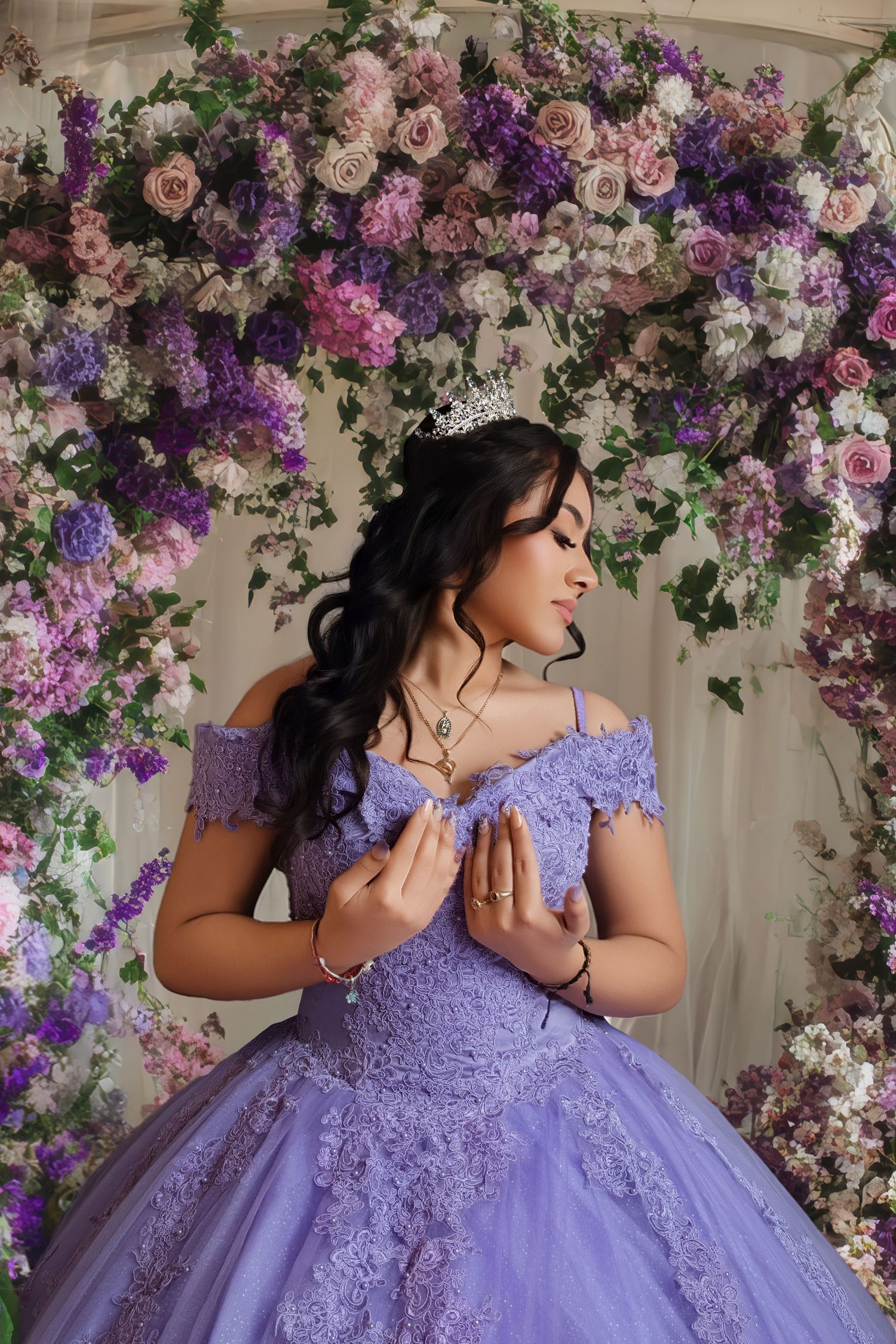 Young woman in lavender quinceañera ball gown with lace, tiara, and floral arch backdrop of purple and pink roses.