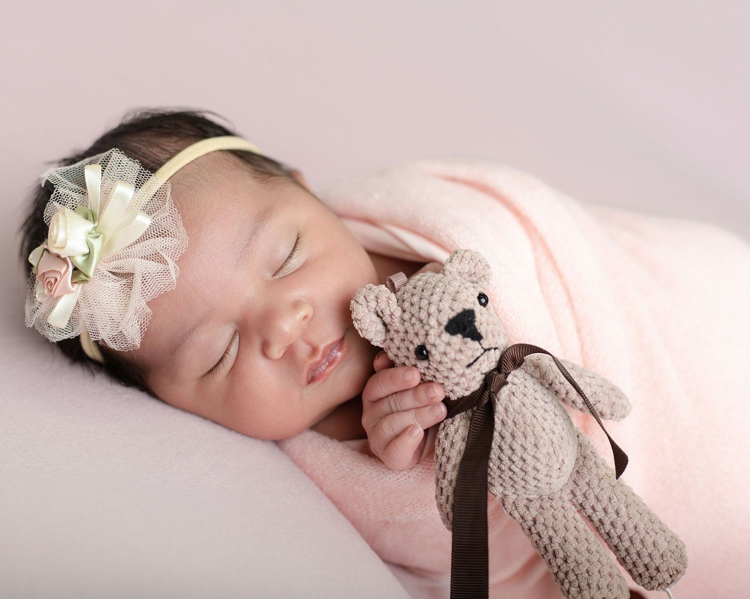 Sleeping newborn baby girl in pink wrap holding knit teddy bear, wearing floral headband, studio photography.